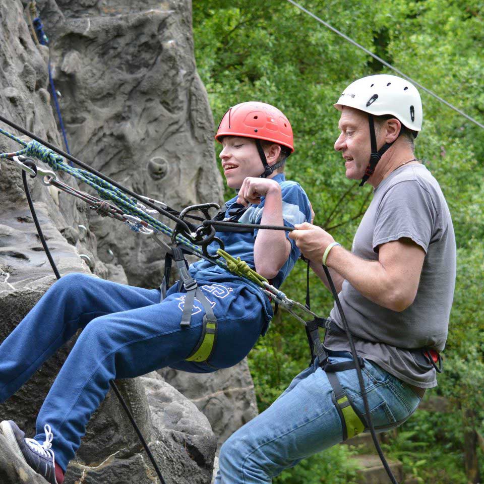 Man and boy tandem abseiling down an outdoor climbing wall with harnesses, ropes and safety equipment, smiling