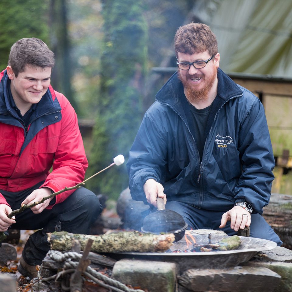 Two male Calvert Trust Exmoor activity instructors smiling, sitting by a campfire cooking food