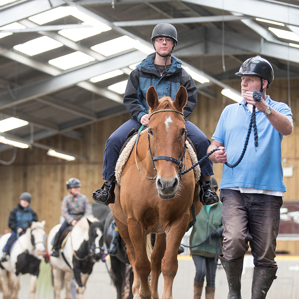 Line of horses being ridden in indoor arena and led by riding volunteers