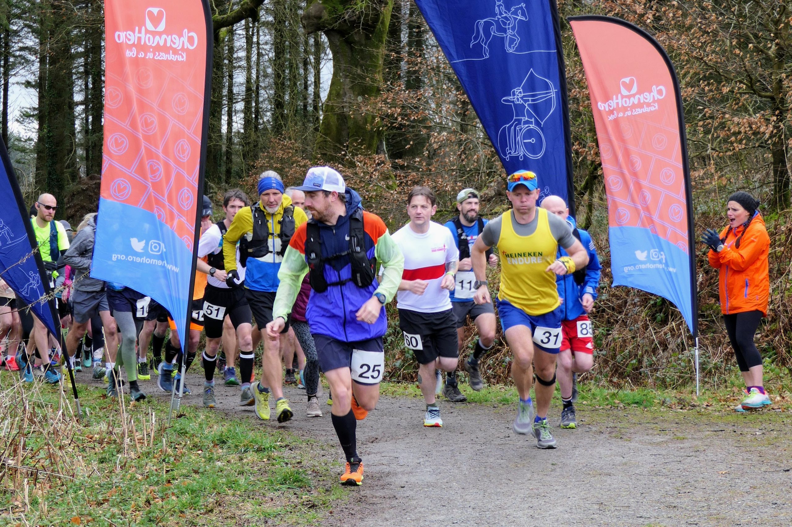 A group of runners running towards the camera on a dirt track in the forrest with Chemohero and Calvert Exmoor flags on the side