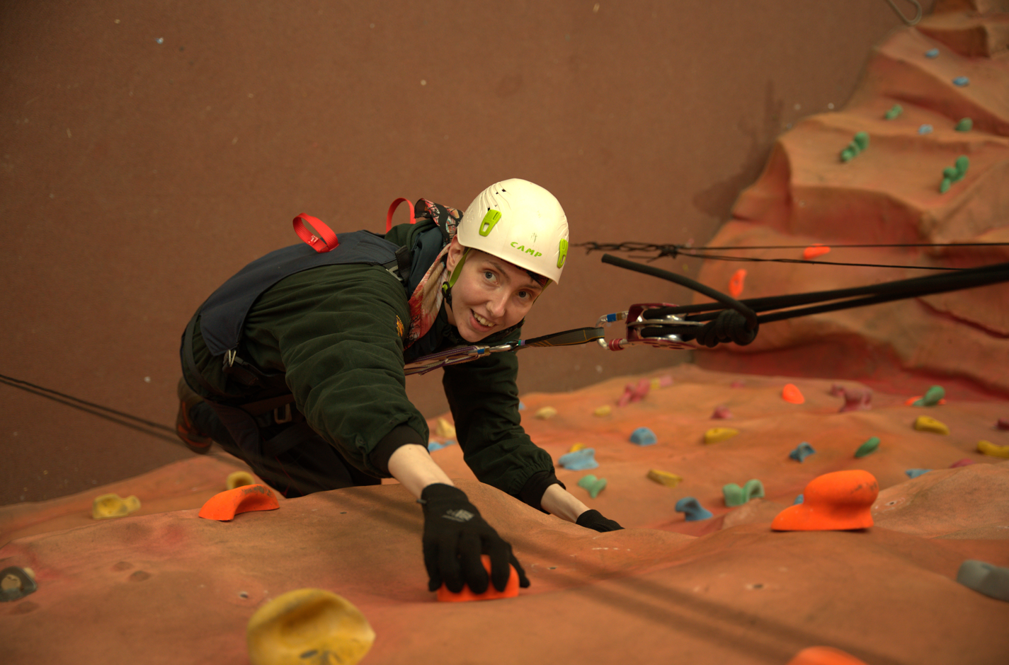A lady in a harness and safety rope system climbing up a wall and smiling at the camera