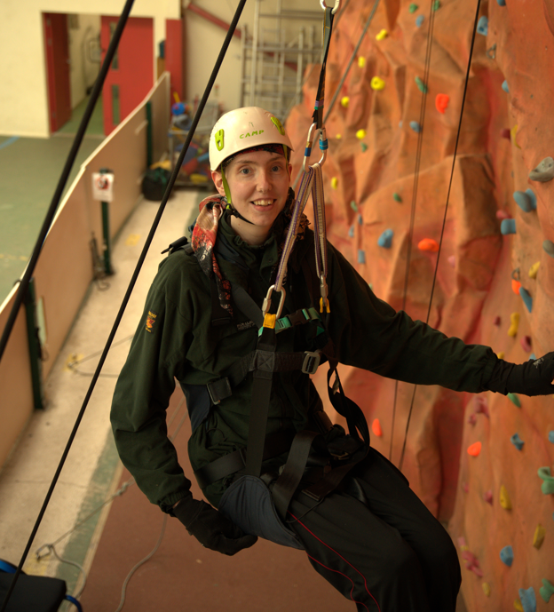 A lady in a harness and safety rope system hanging next to a climbing wall and smiling at the camera