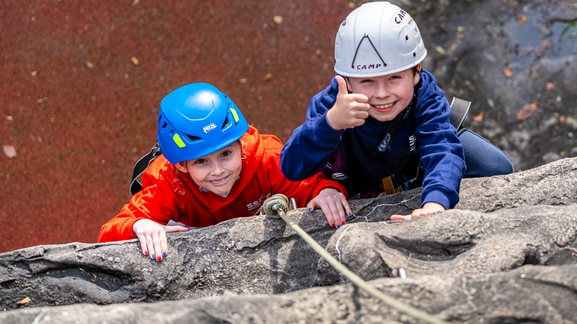 Two children climbing a rock wall with safety rope, one is giving a thumbs up to the camera