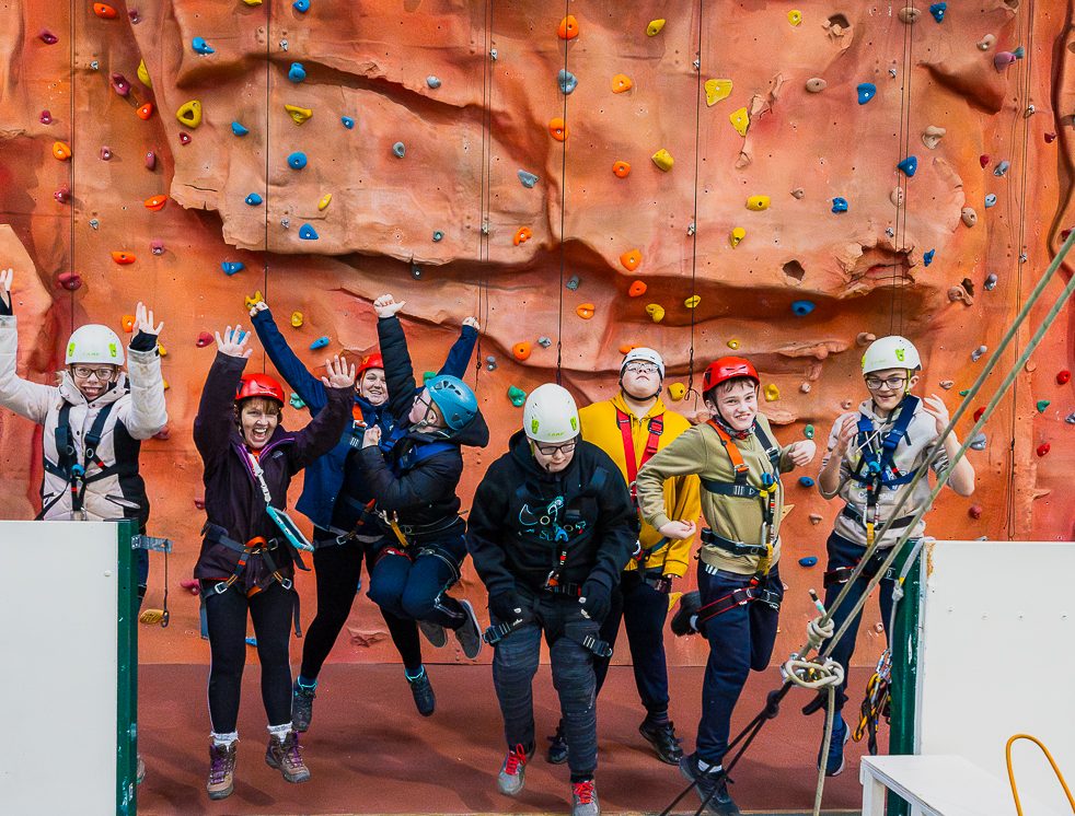 A group of children and adults jumping into the air in their climbing gear and helmets in front of an orange climbing wall