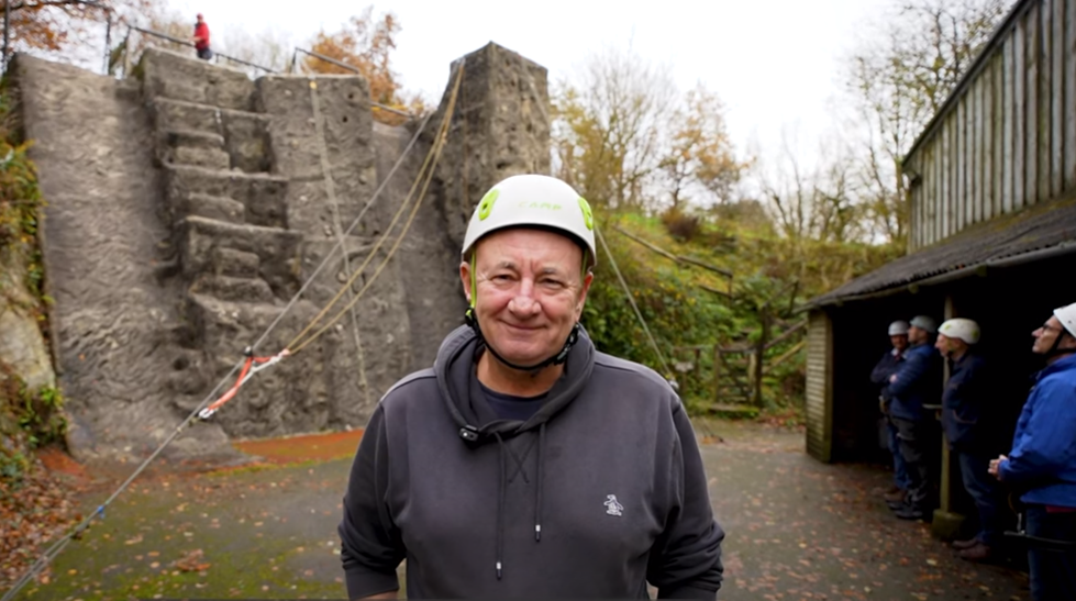 A man in a white climbing hat stood infront of the camera and looking straight at it with an outdoor climbing wall in the background