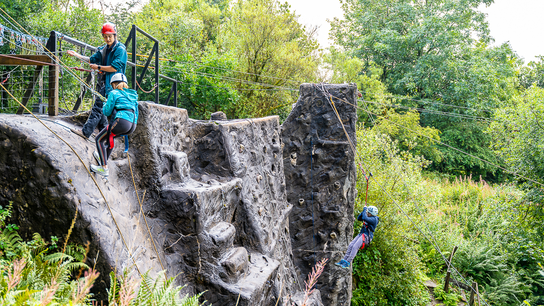 A Calvert Exmoor instructor is at the top of the abseiling wall looking at a young girl who is starting her abseiling activity and is slightly further down on the wall. They are both attached into a rope system and in the background there is a child being hoisted up the climbing wire to the top of the climbing wall on a harness.