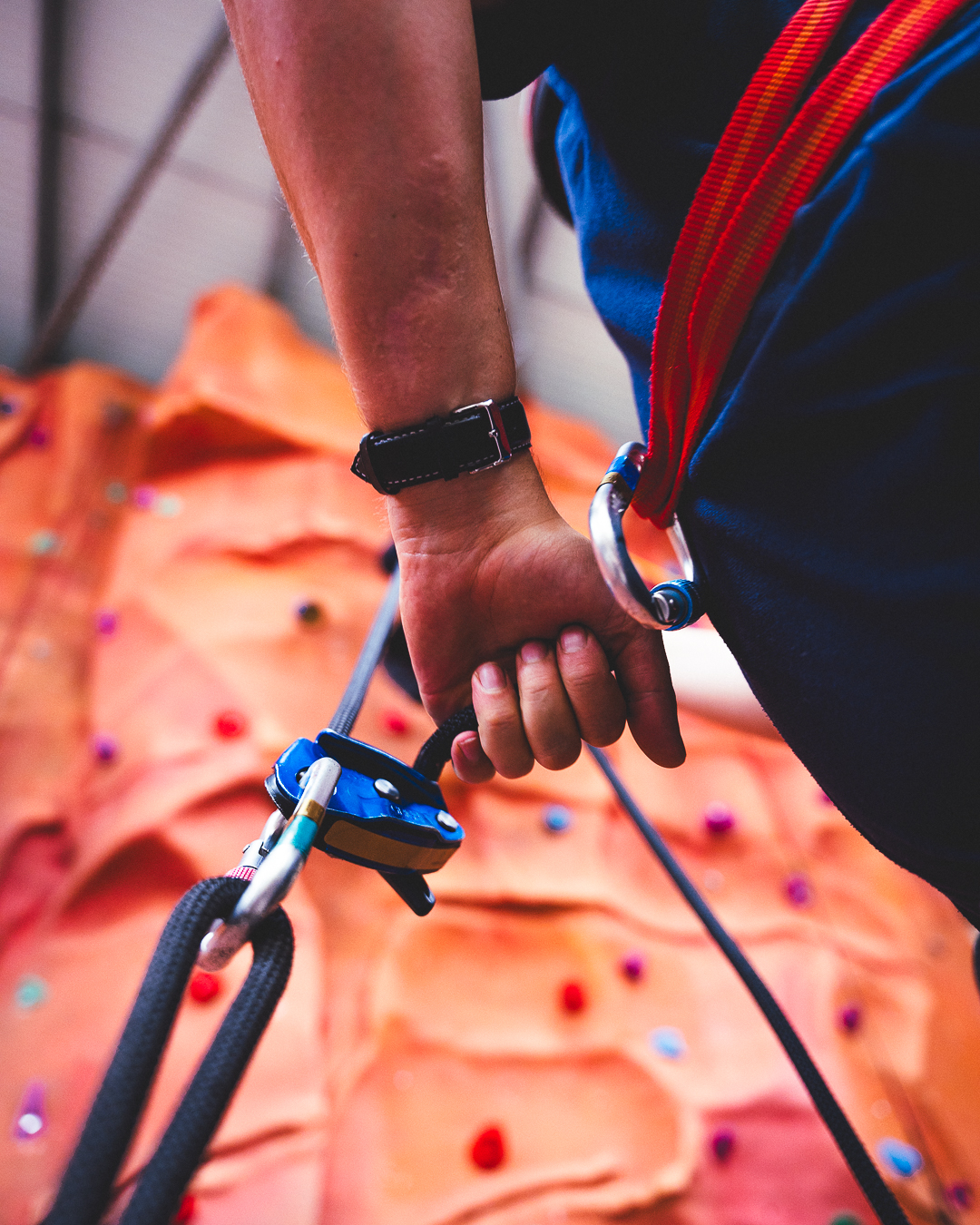 The camera is at knee-height and facing upwards, focused on a piece of climbing equipment and a person's hand and waist next to the equipment with the orange indoor climbing wall behind.