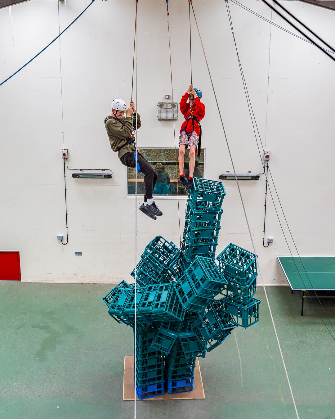 Two people with harnesses and helmets on are suspended high in the air on ropes with a tower of crates beneath them crumbling to the floor.