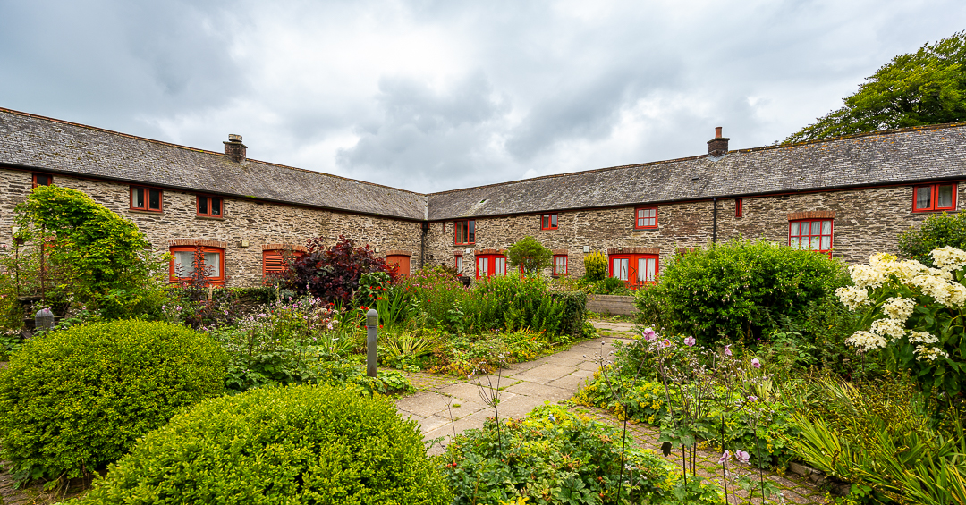 The courtyard in the middle of the accommodation buildings of Calvert Exmoor. There is beautiful foliage such as bushes, plants, flowers and trees in the foreground and in the background, stone built wall and red window frames and door frames of the accommodation buildings.