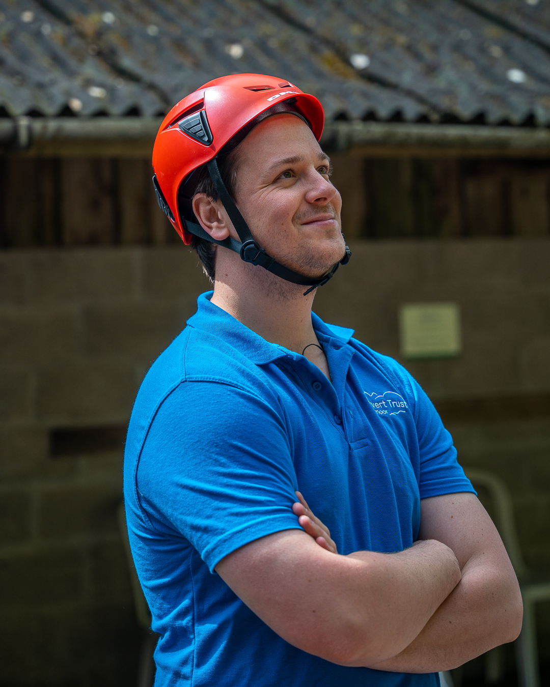 A man wearing a light blue Calvert Exmoor volunteering polo shirt and a red helmet has his arms crossed and his looking upwards and smiling at something out of frame.