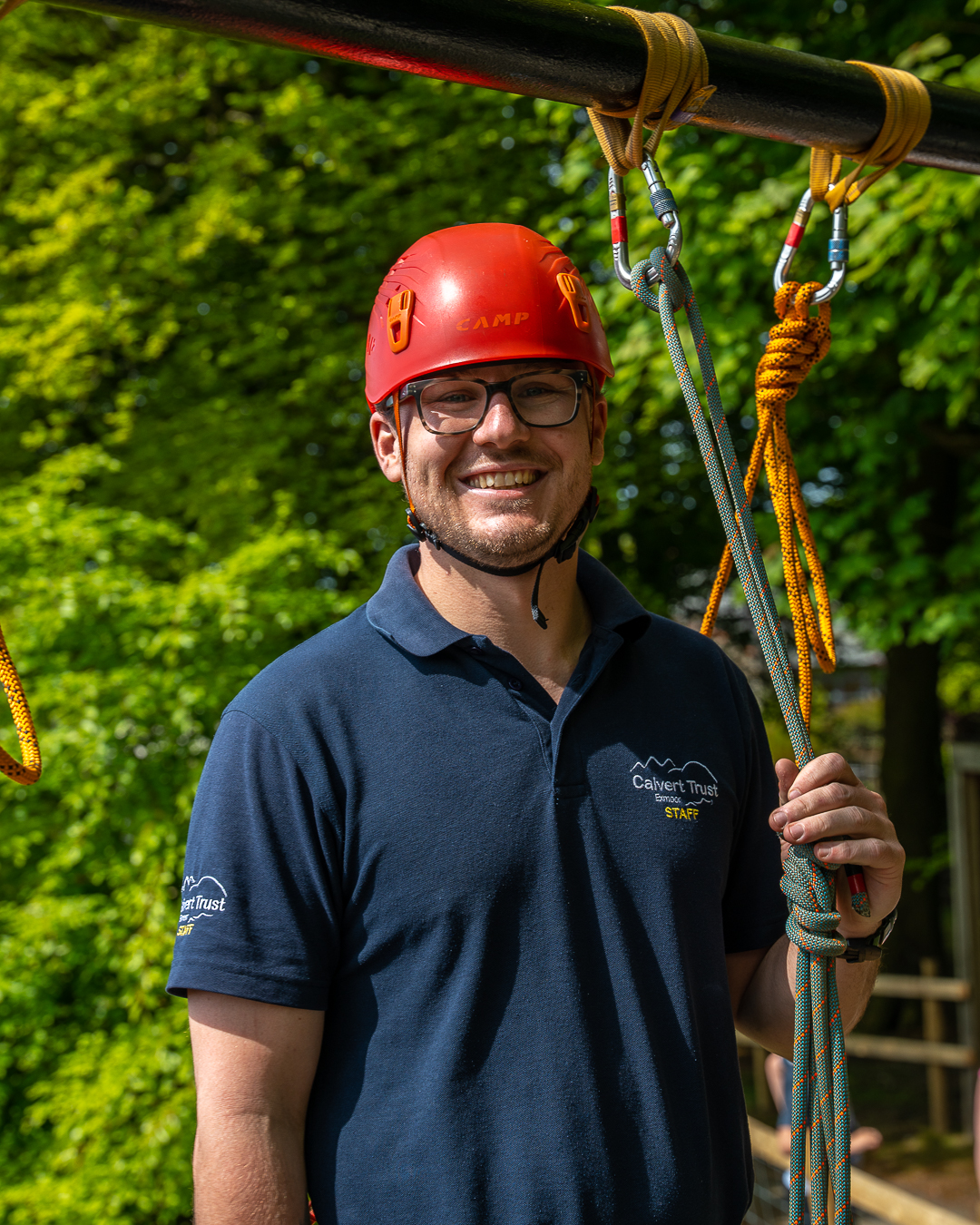 A member of Calvert Exmoor staff, Chief Instructor Tom, is wearing a navy blue Calvert polo shirt, a red helmet and glasses and is holding some ropes tied to a carabiner, smiling at the camera.