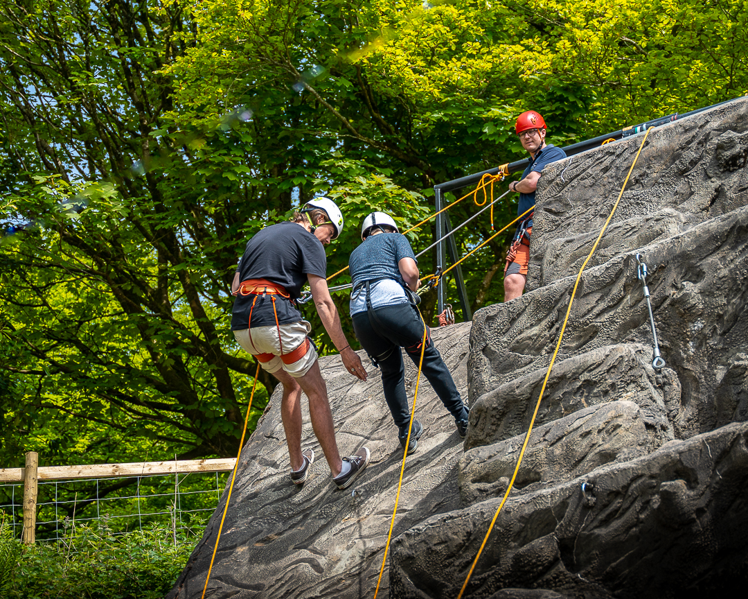 An instructor at the top of the abseiling wall looks down at two guests facing away from the camera descending down the climbing wall next to each other, all attached by ropes and harnesses.