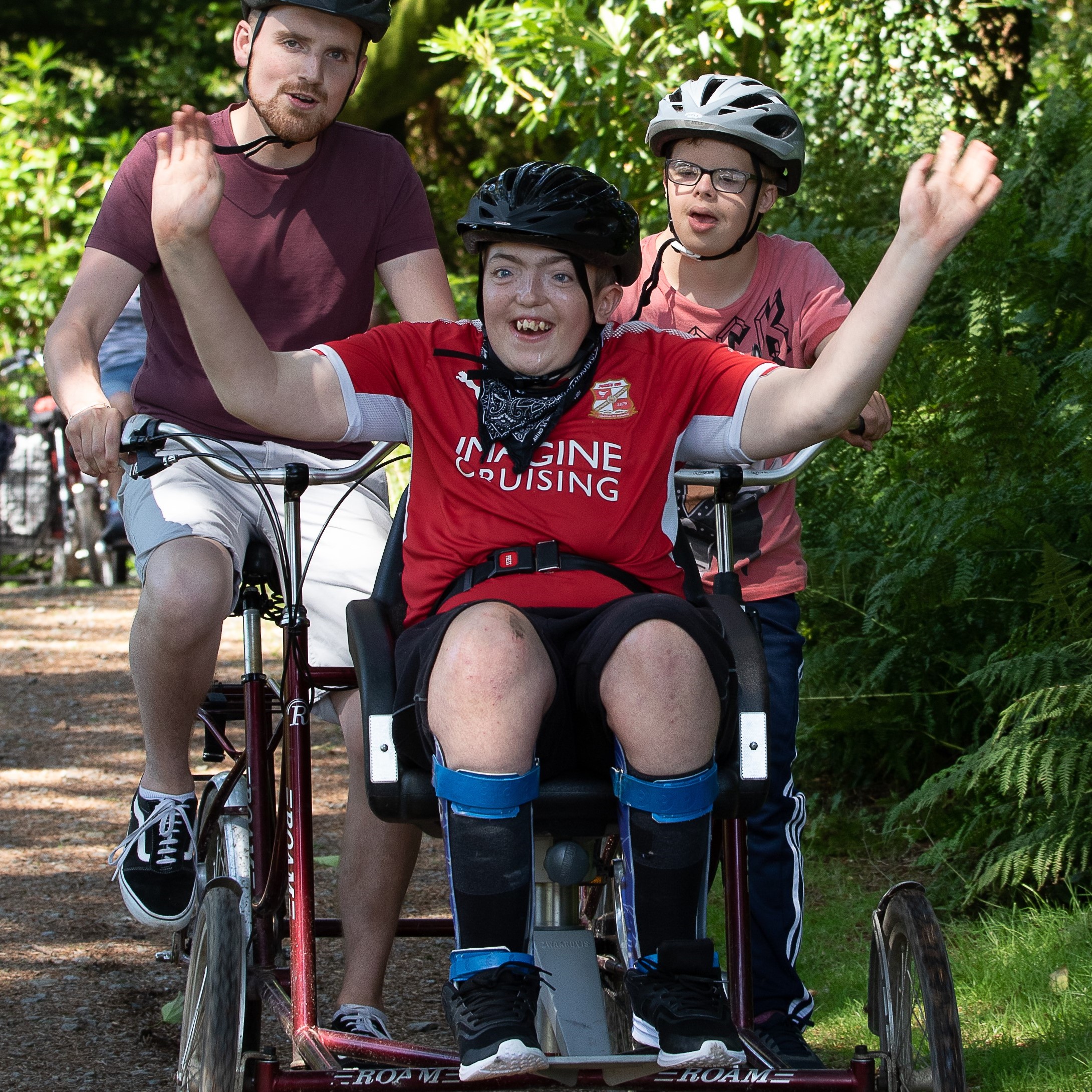 Zoomed in picture of a guest wearing a black helmet and a red t-shirt is sitting on a chair on the front of a four-wheeled bike that has two people sitting and pedaling on behind. They are all looking towards the camera and the guest at the front is smiling with his hands in the air.