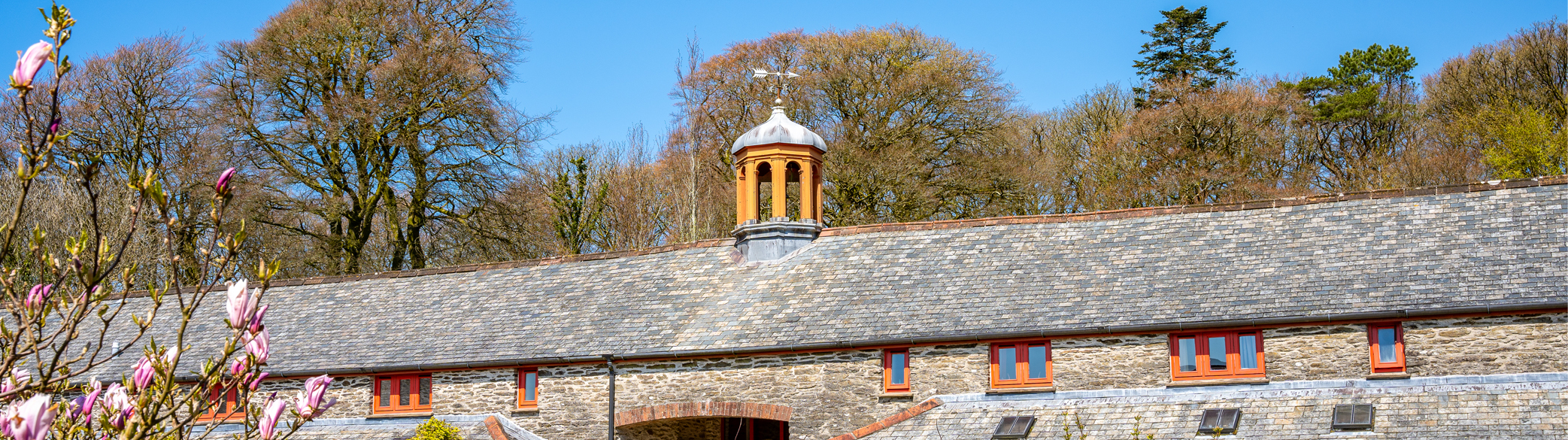 A wide-angled photograph of the long stone building of Calvert Exmoor accommodation, with a belltower atop the roof. The windows are all red-framed and there is a blooming magnolia tree in the bottom left of the photograph.