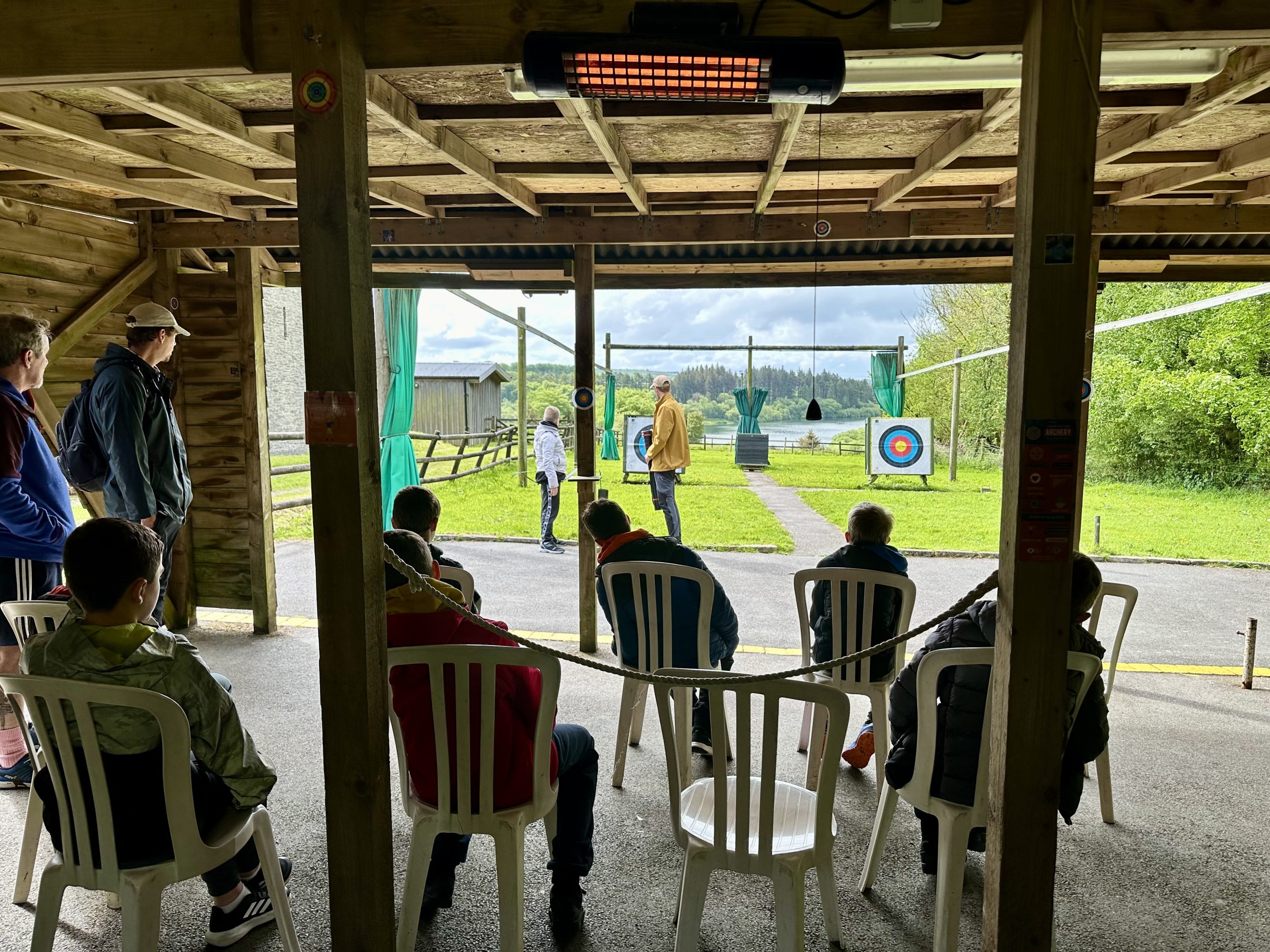 A collection of white plastic chairs have young guests sitting facing away from the camera, looking out at the archery range in the background, where a Calvert Exmoor instructor is teaching a young guest how to shoot and arrow with a bow.
