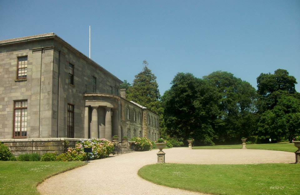 A tall grey building with pillars stands on the left, with a clear blue sky behind and a paved area in front of the house surrounded by a tidy lawn.