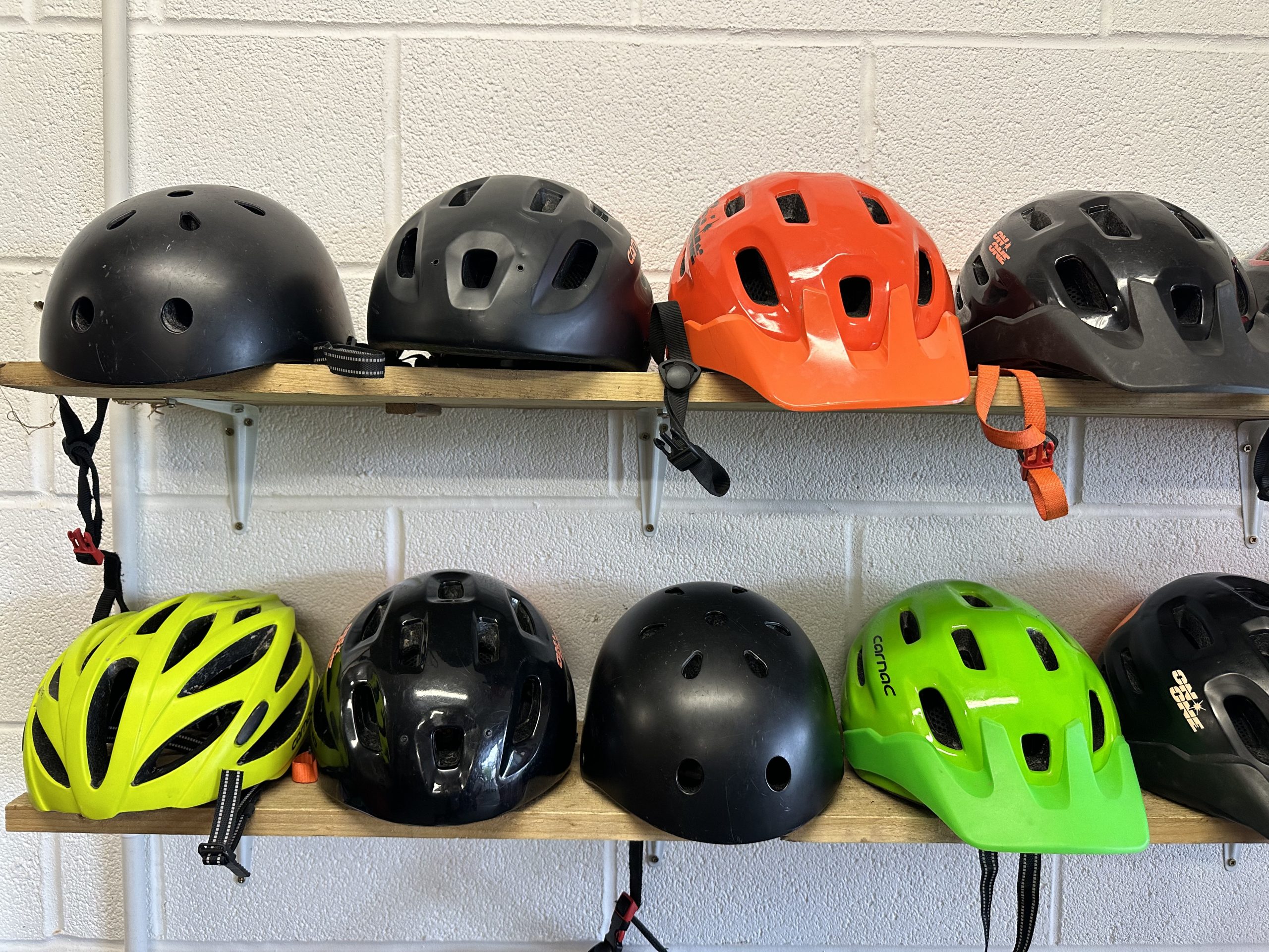 A row of bike helmets are lined up on two shelves. There are black, orange, green and yellow helmets.