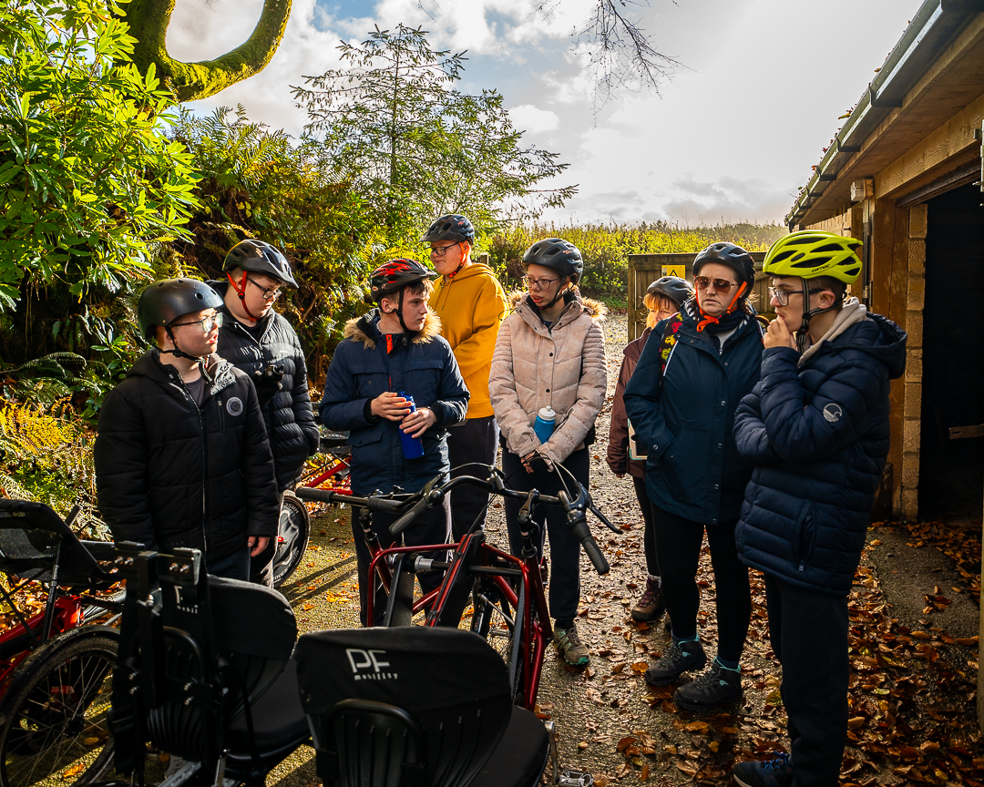 A group of guests are wearing biking helmets in a circle, looking at the selection of bikes they can use for their accessible biking session. On the right is the bike shed made of wood paneling.