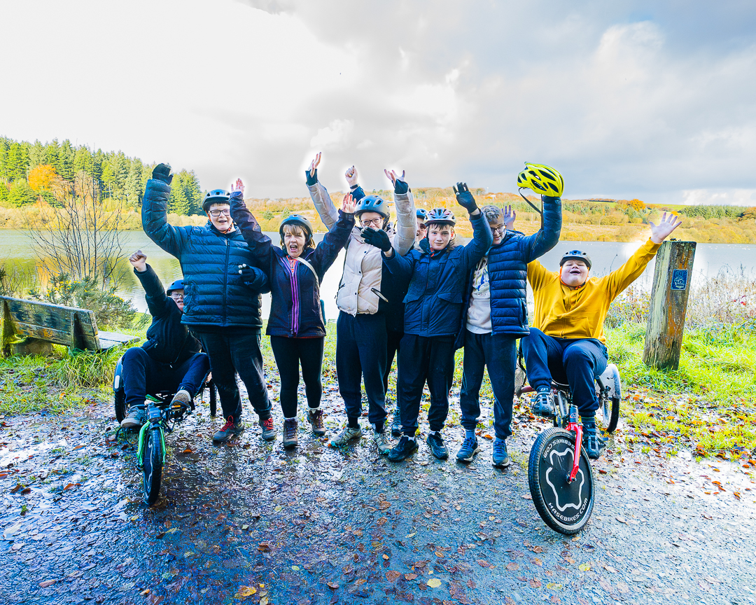 A group of guests wearing helmets or lifting helmets and their arms into the air in front of Wistlandpound Reservoir. Two guests either side are sitting on their bikes and the others are standing in between.
