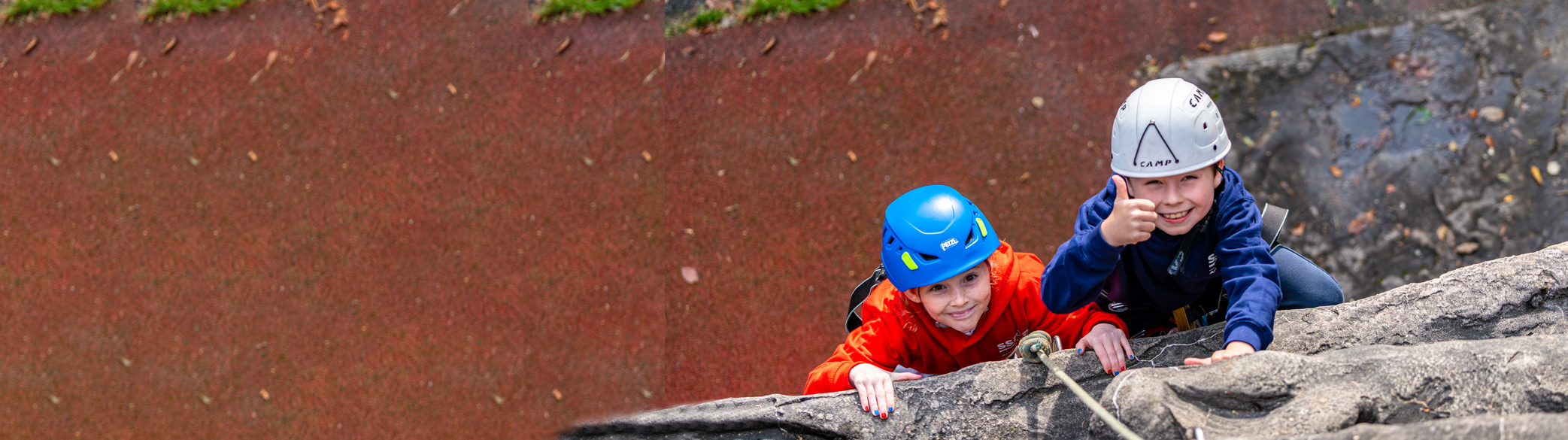 A wide-angled photograph from aerial view down the outdoor climbing wall of two boys climbing upwards and smiling towards the camera. One boy has his thumb up at the camera too.