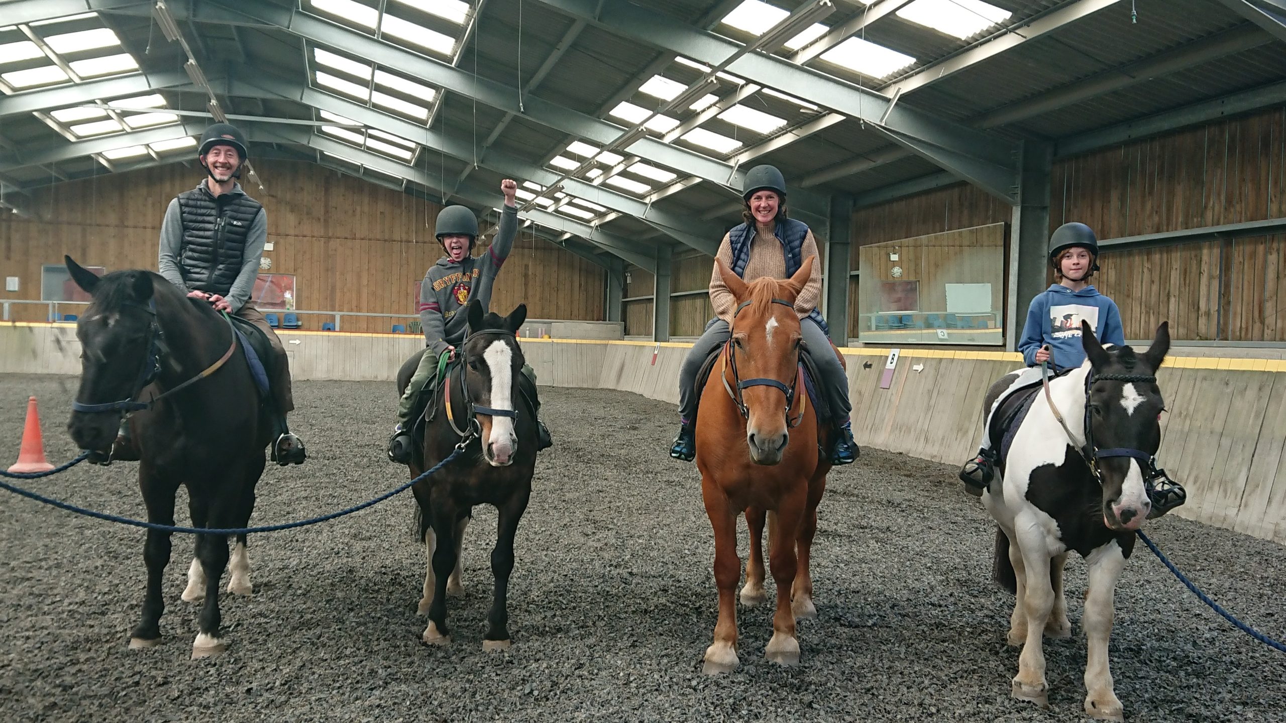 Four people sit on top of a horse each in a line facing the camera. They are all wearing riding hats and smiling in an indoor arena building.