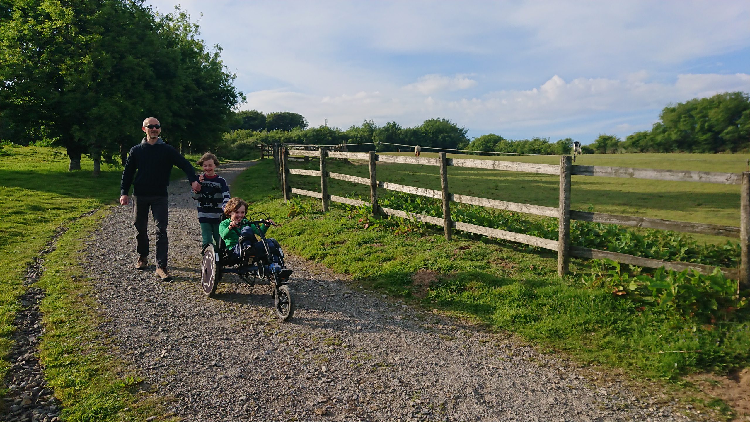 A man wearing sunglasses is holding the arm of a young boy to the right and they are smiling walking downhill behind a boy who is on a recumbent bicycle, smiling and cycling.