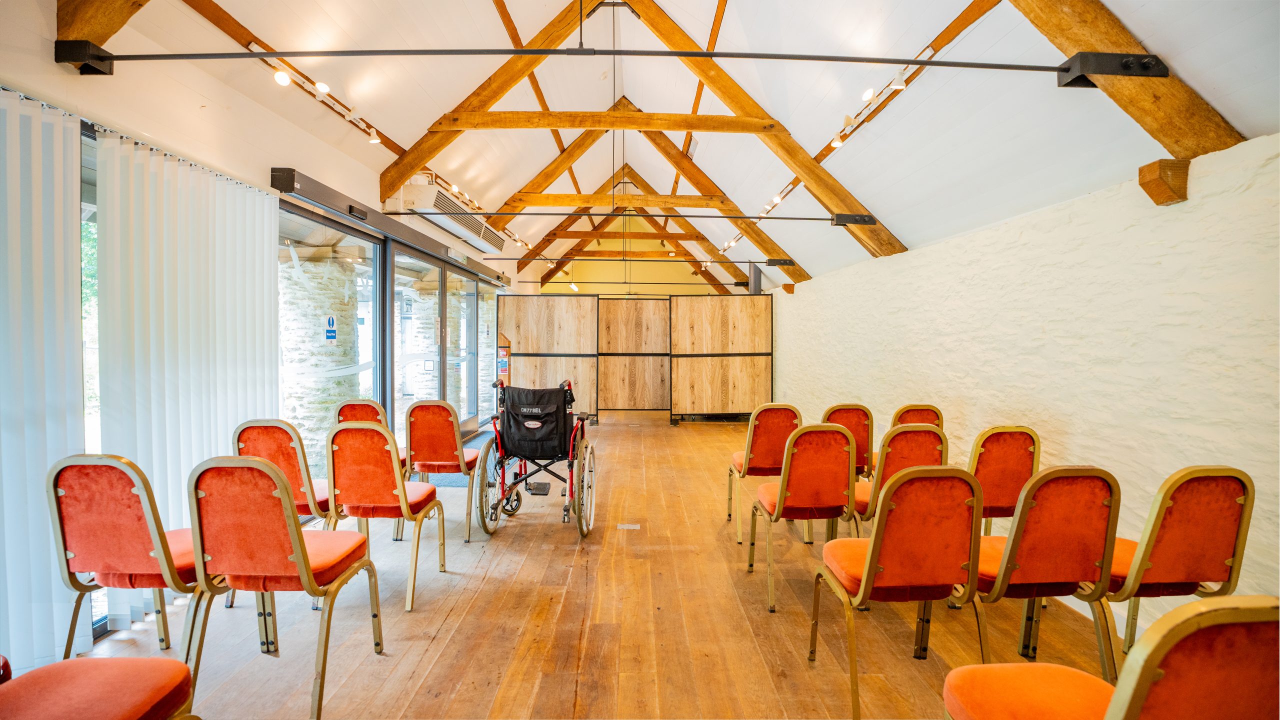 A row of orange chairs face away from the camera, with an aisle of empty space in the middle. There's ablack wheelchair among the chairs. There are windows on the left with blinds and a white coloured wall on the right, with wooden beams above in a triangle shape up to the roof.