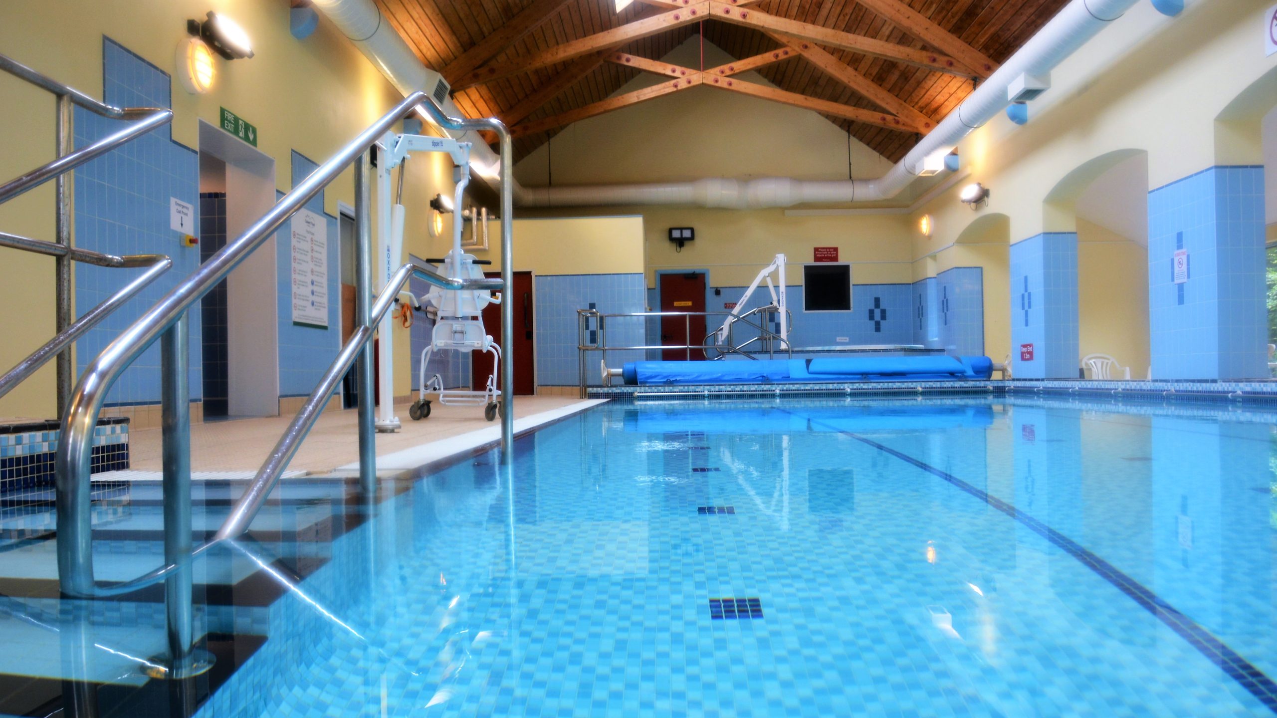 An empty indoor swimming pool with blue tiling and yellow walls, with a wooden paneled ceiling and wood beams. There are silver railings in the foreground with steps that go into the pool and a hoist in the background.