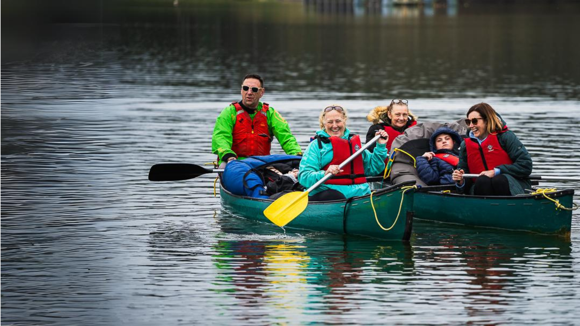A group of 6 people are smiling and enjoying canoeing on the calm water. The two canoes are rafted together and there is a guest in a beanbag in the middle of each one.