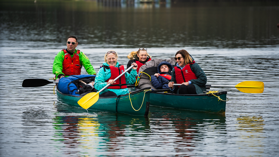 A group of guests are sitting in a pair of rafted canoes all laughing and smiling, wearing red buoyancy aids and paddling on lightly rippling water. There are two guests sitting at the front, two guests sitting at the back and then the guests in the middle of each boat are in beanbags.