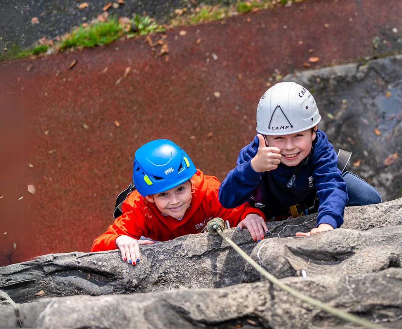 An aerial view of two boys climbing the outdoor climbing wall wearing helmets and smiling at the camera. The boy on the right is giving a thumbs up to the camera.