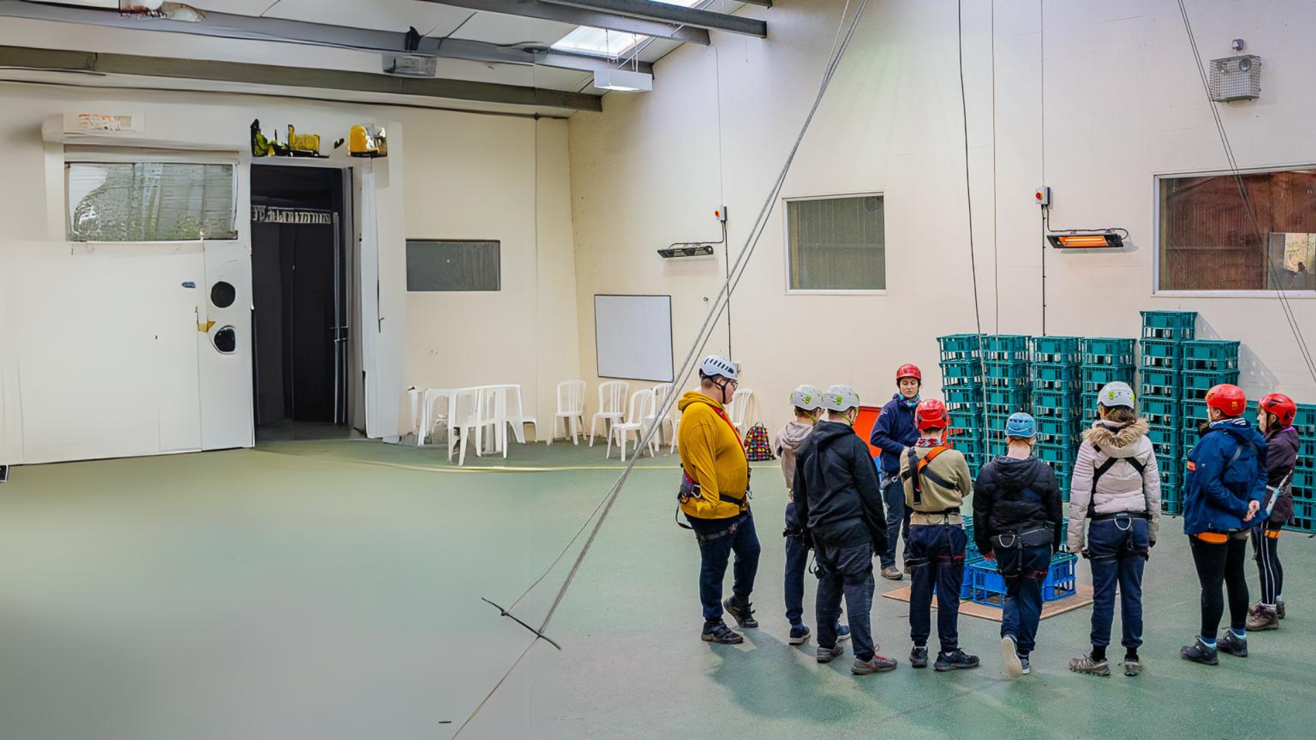 An extended view of the indoor sports hall at Calvert Exmoor. To the left is empty space and to the right is a group of people wearing harnesses and helmets with their backs to the camera, watching a Calvert Exmoor instructor who is explaining the activity of Crate Stacking. There is a pile of crates in the background.