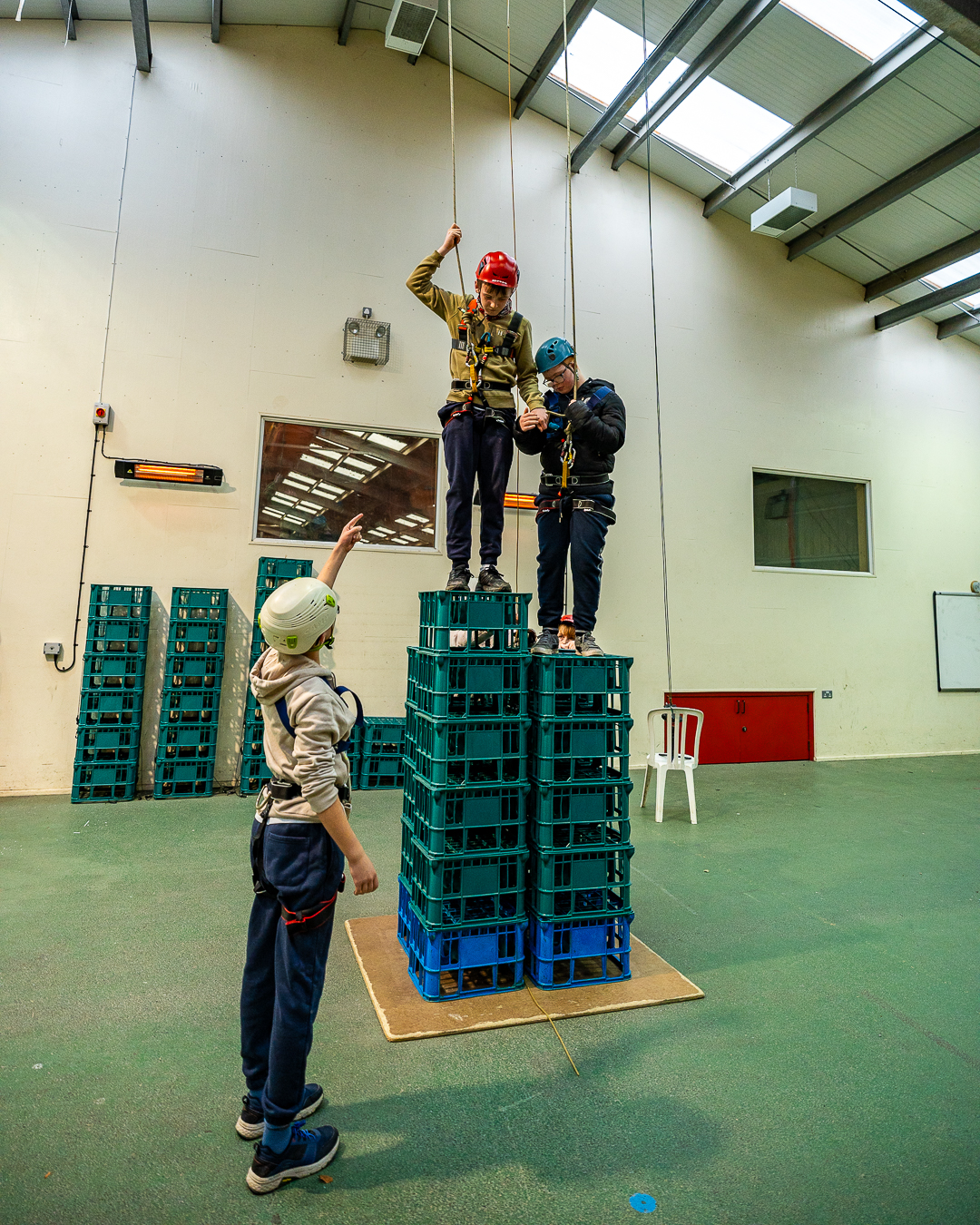 Two guests are standing on top of a tower of crates, attached by harnesses and ropes to the ceiling out of shot and they are looking down at a person standing on the floor who is pointing and directing them what to do.
