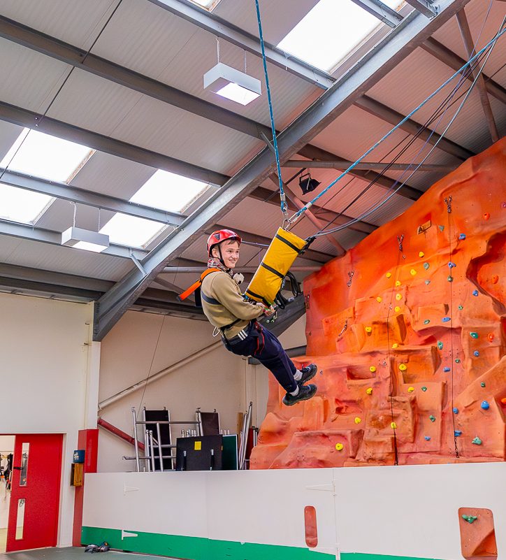 A guest is suspended in the air on a rope system in the indoor sports hall, with the climbing hall in the background. They're smiling as they're swinging through the air on the Giant Swing.