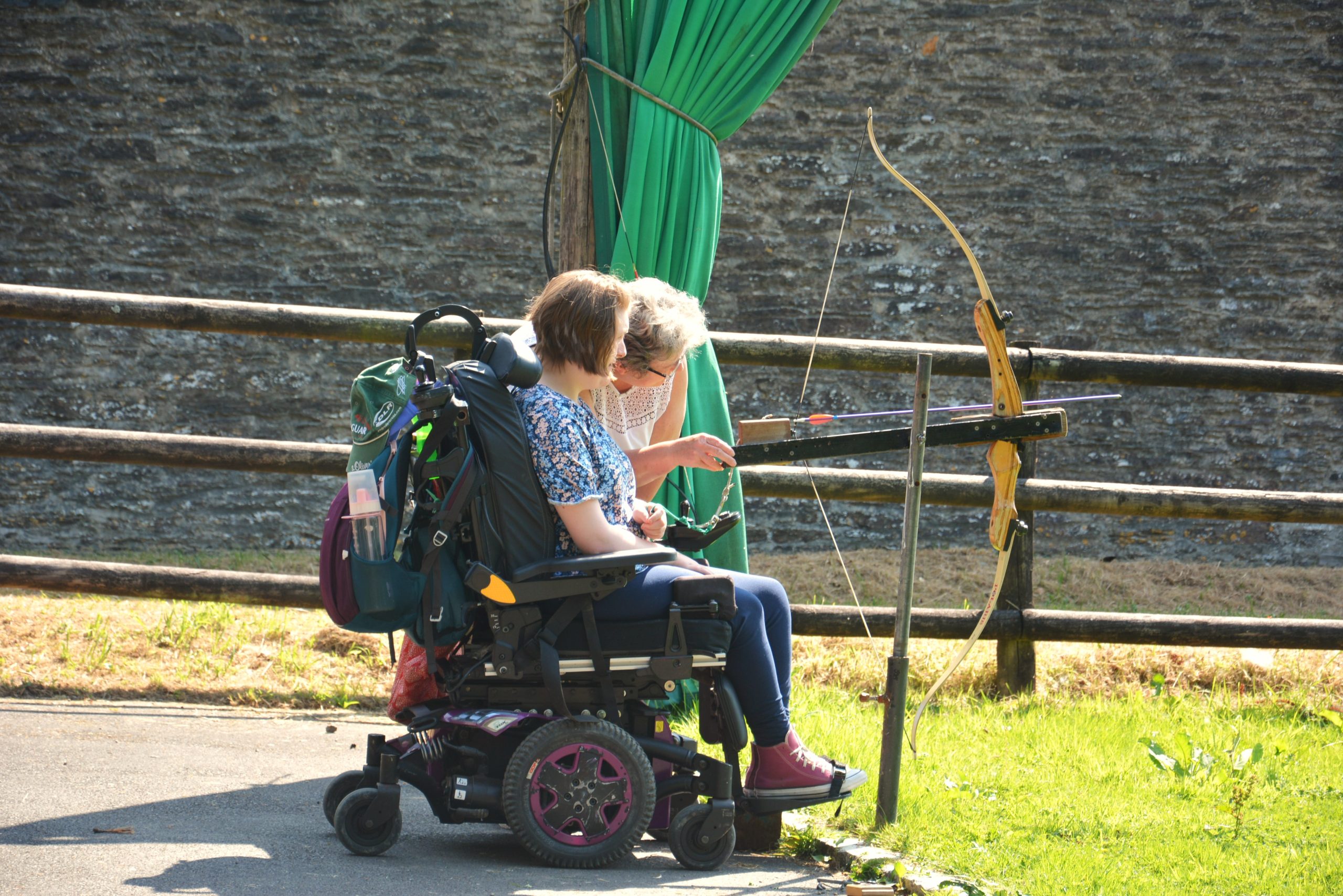 A guest in a wheelchair is looking to the side, with another guest leaning down to help them use the accessible trigger bow. The bow is loaded and ready to fire, pointing to the right.