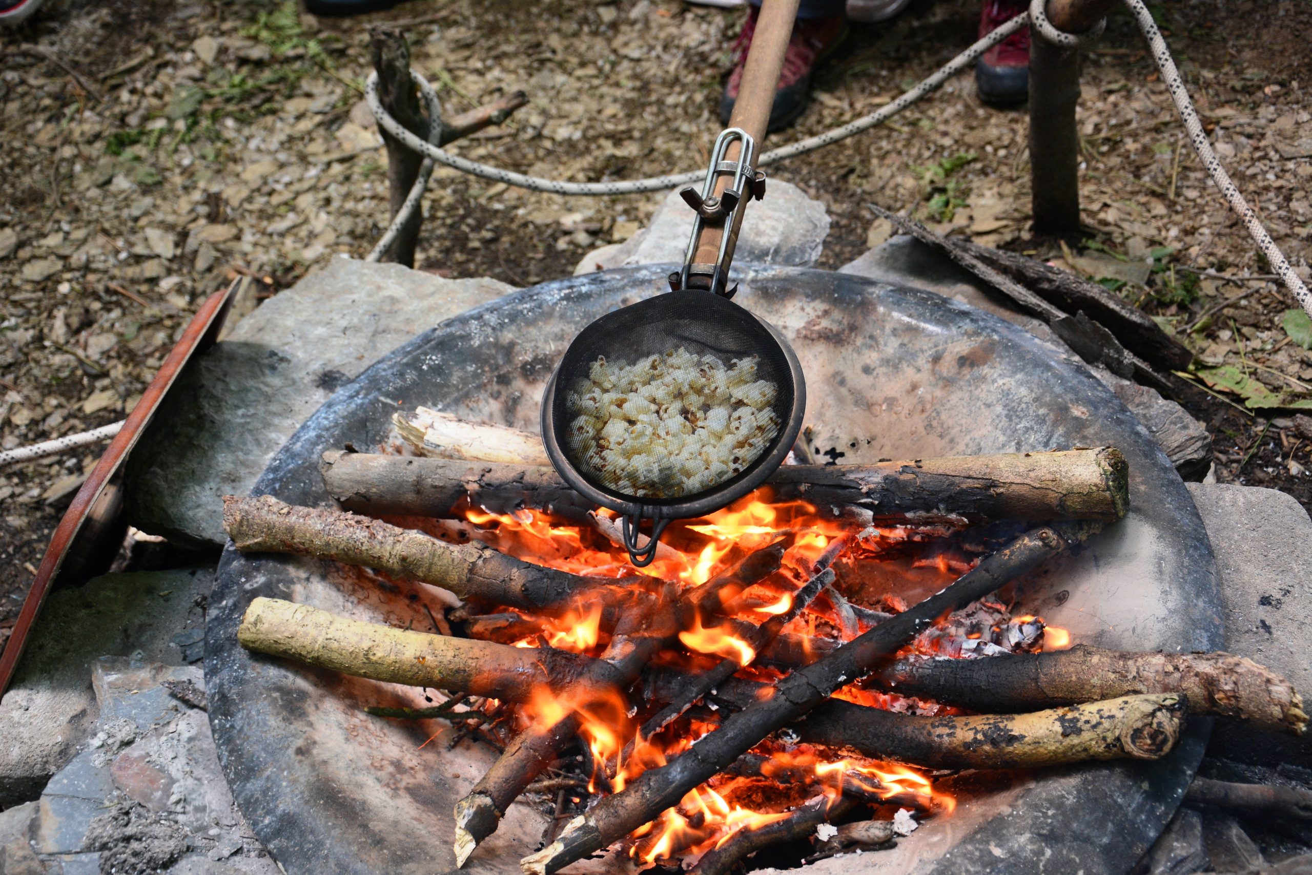 A contraption that consists of two sieves on top of each other (to create a bubble shape) is over a fire with popcorn inside it. There is wood in the firebowl, ropes in the background and dead leaves and twigs all over the floor.