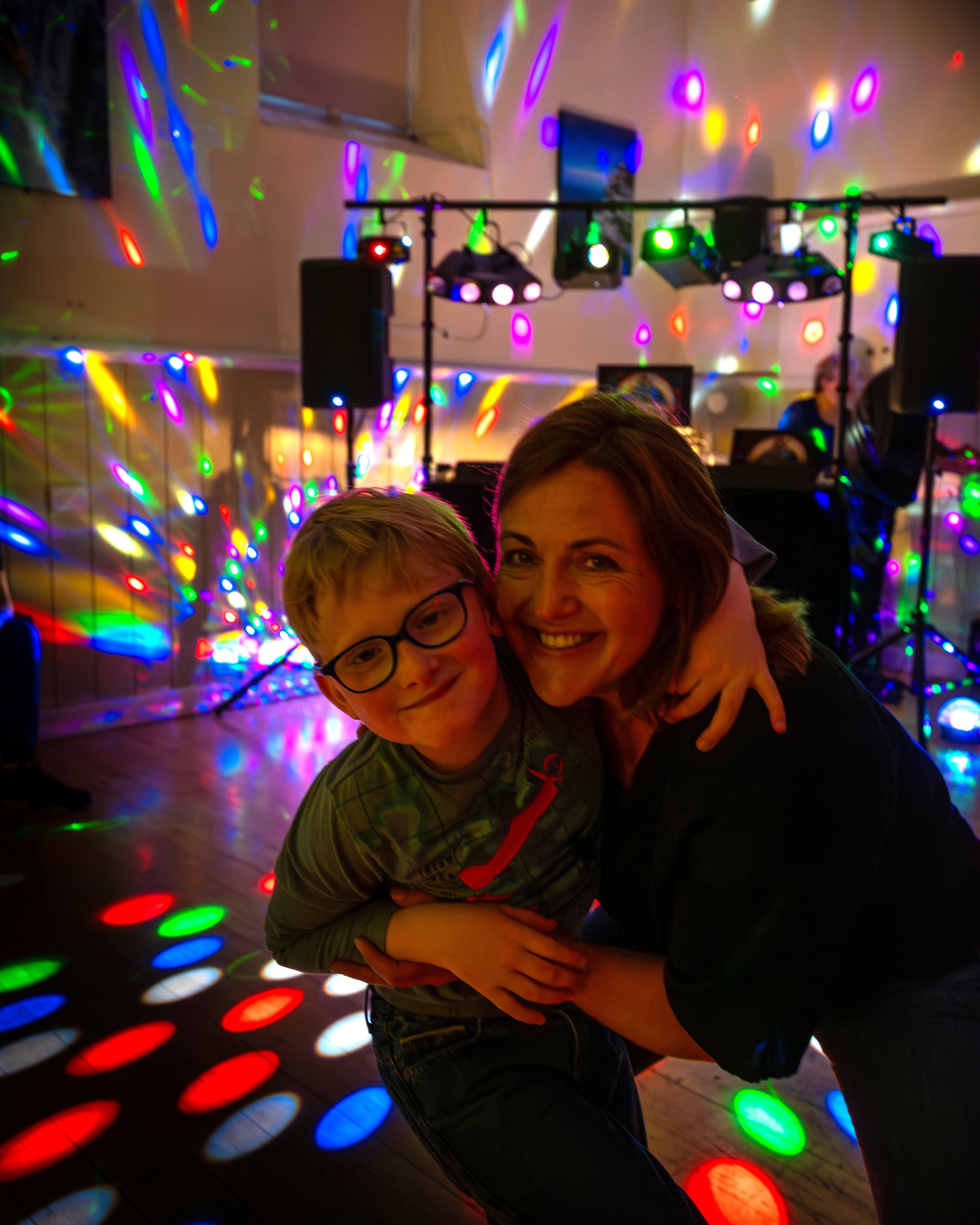A DJ deck with speakers and lights in the background, with a woman and a young boy wearing glasses smiling at the camera in the foreground.