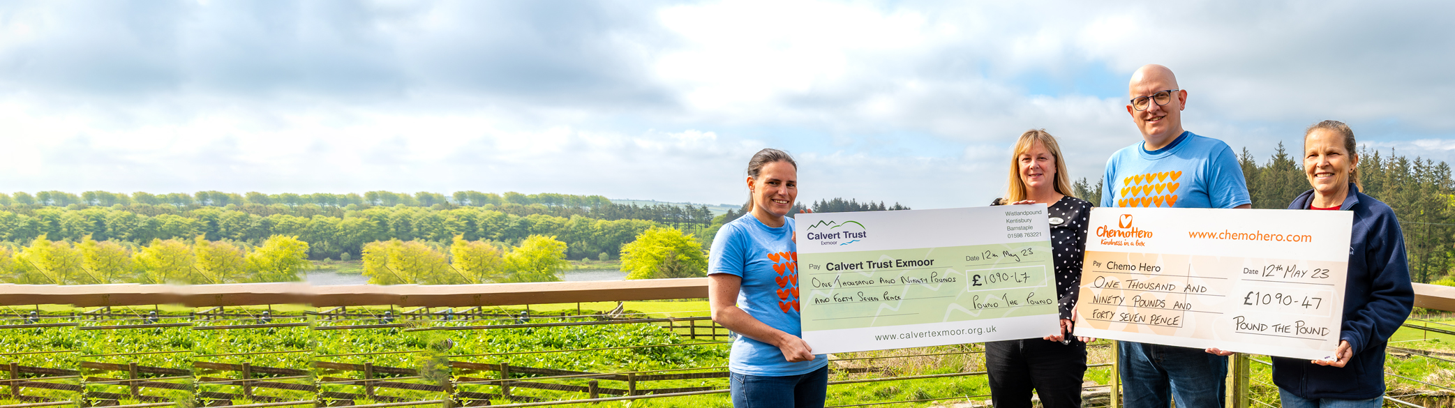 A wide-angled photograph of the balcony with the view of a reservoir behind. To the right is four people, each pair holding a large cheque. One cheque is made out to Calvert Exmoor and the other is made out to Chemo Hero. All four are smiling at the camera in the sun.
