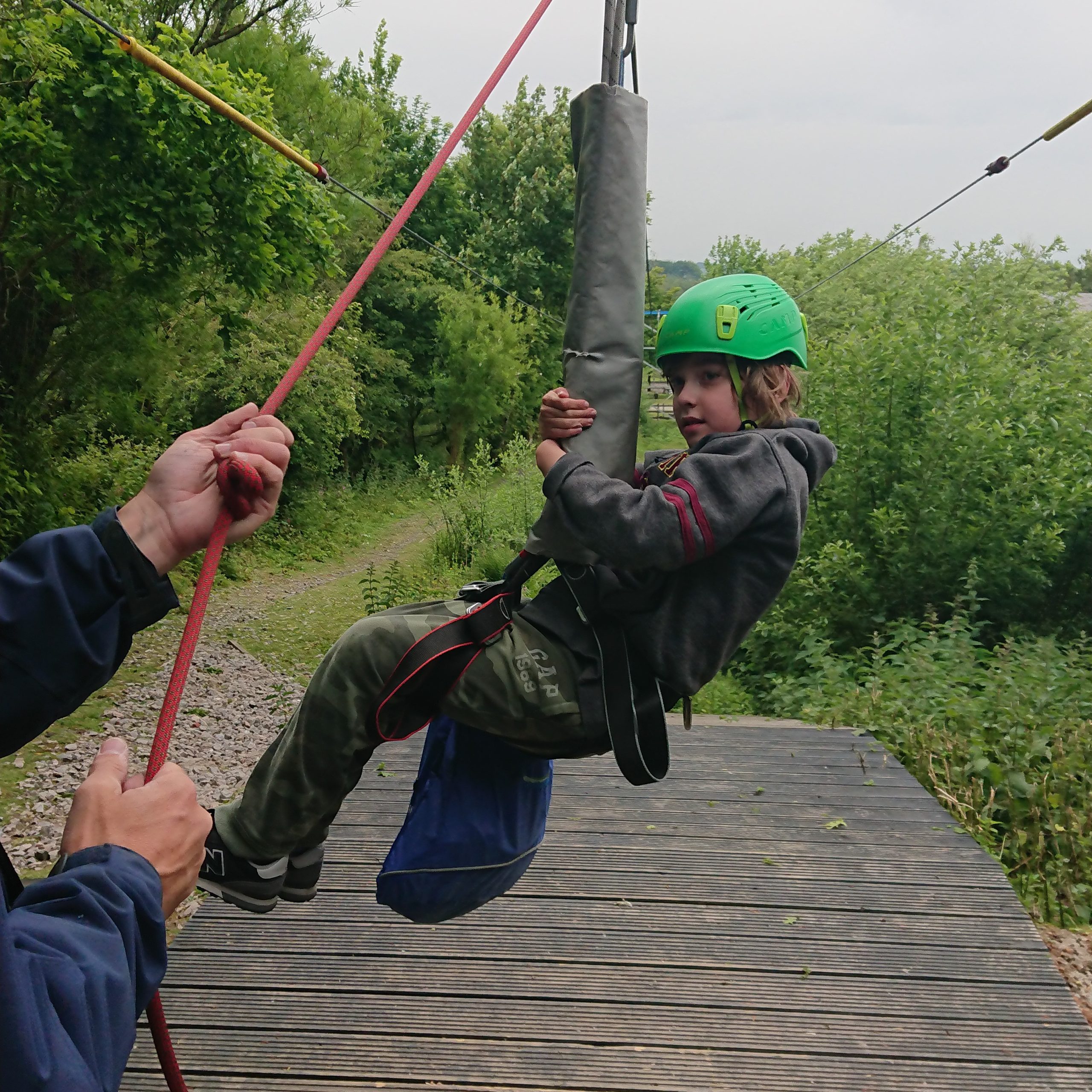 A boy wearing a dark green sweatshirt and tracksuits and a bright green helmet is looking at someone whose arm is in the photo, holding a red rope. The boy with the green hat is suspended in a seated position in the air by a zipwire and harness.