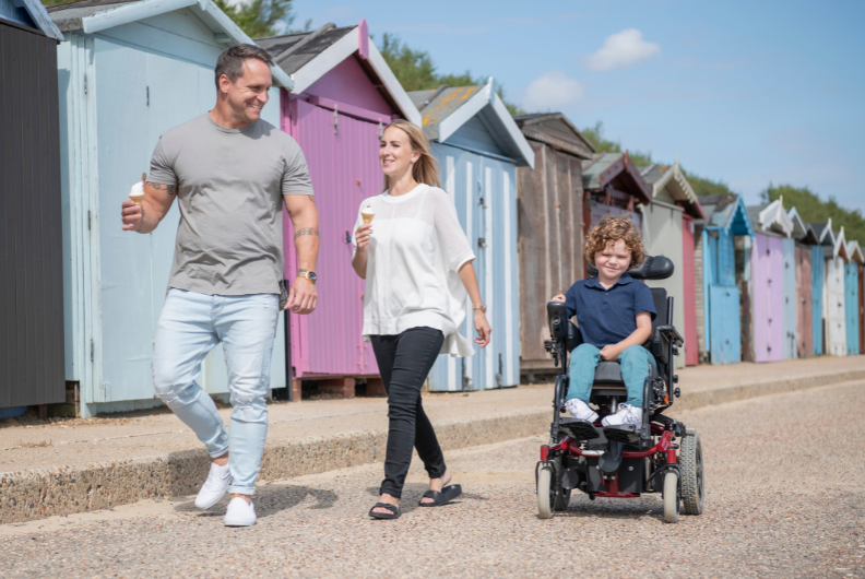 A man holding an ice cream is walking and looking back at a young boy in a powered wheelchair. A woman is walking between the two of them and in the background there are colourful beach huts.