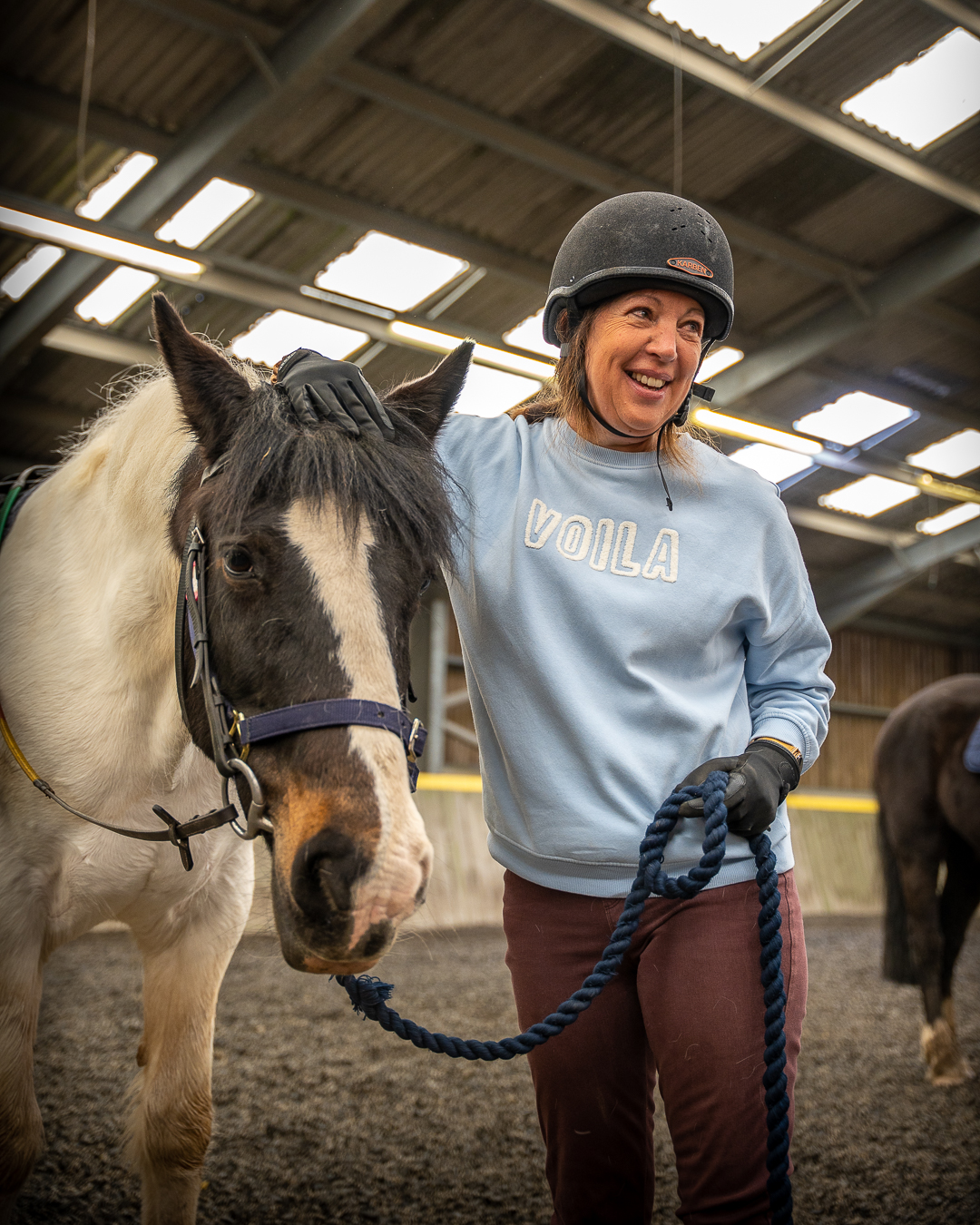 A woman wearing a light blue sweatshirt, holding the lead rope for a brown and white horse and wearing a riding had is standing and smiling looking away from the camera in the indoor riding arena at Calvert Exmoor.