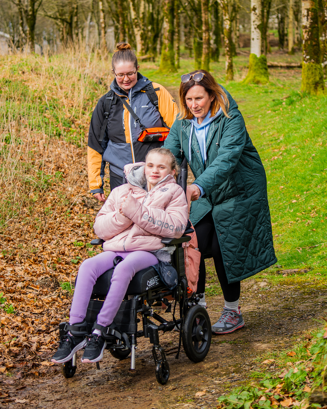 A guest in a wheelchair is being pushed down the hill on a paved path by another guest in a long green coat. Behind them is an instructor walking down the path with greenery either side of them all.