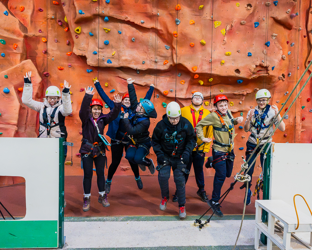 A group of guests are wearing helmets and harnesses and are jumping with joy at the bottom of the indoor climbing wall, facing the camera.