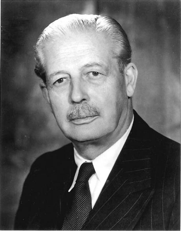 A black and white image of a man in a pinstripe suit. He has slicked back hair and a moustache and is looking at the camera with a serious face.