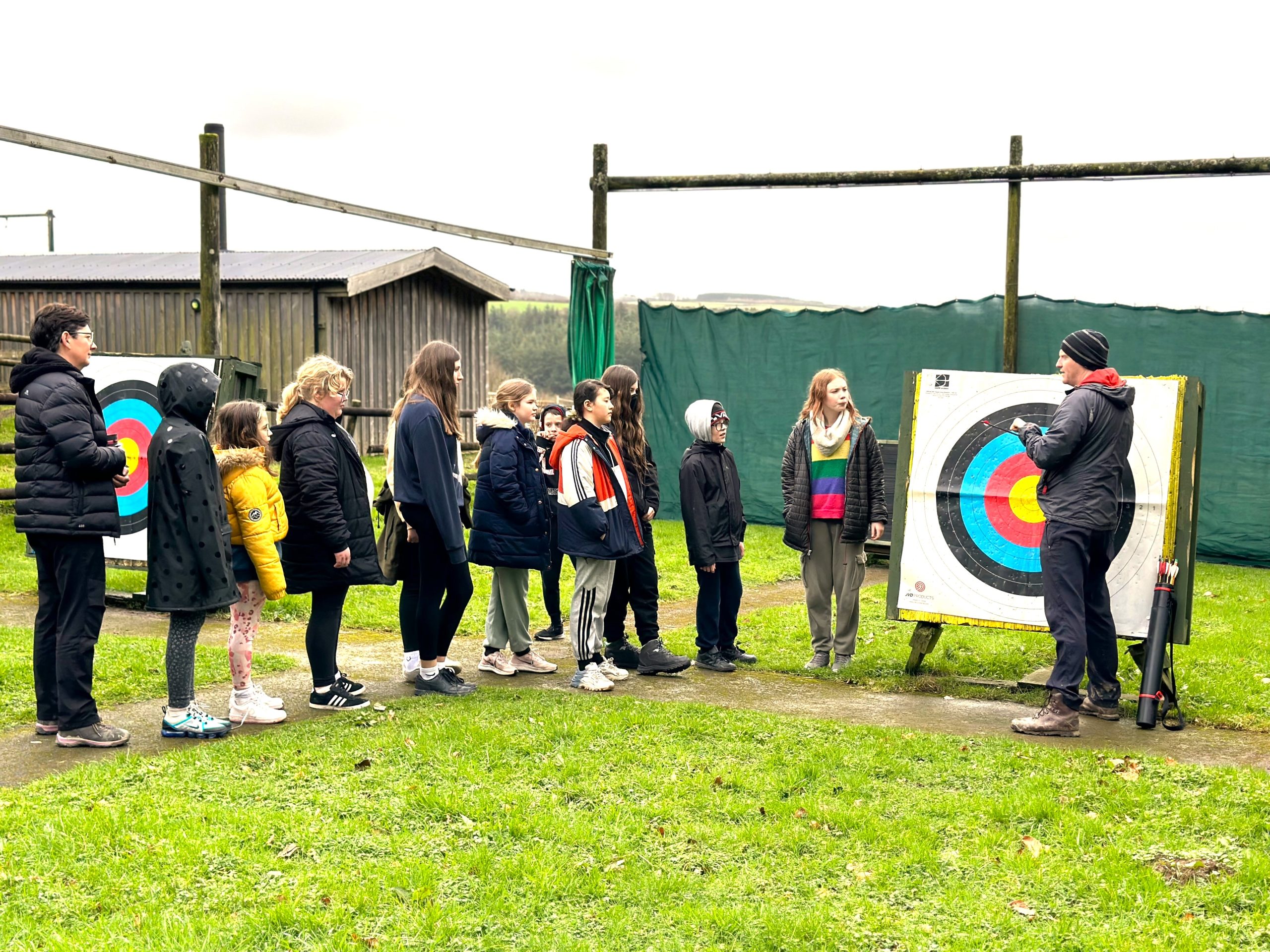 A group of young guests are gathered along the path that leads to the target in the archery range. They are looking at the instructor, who is standing in front of the target, talking to them about archery.