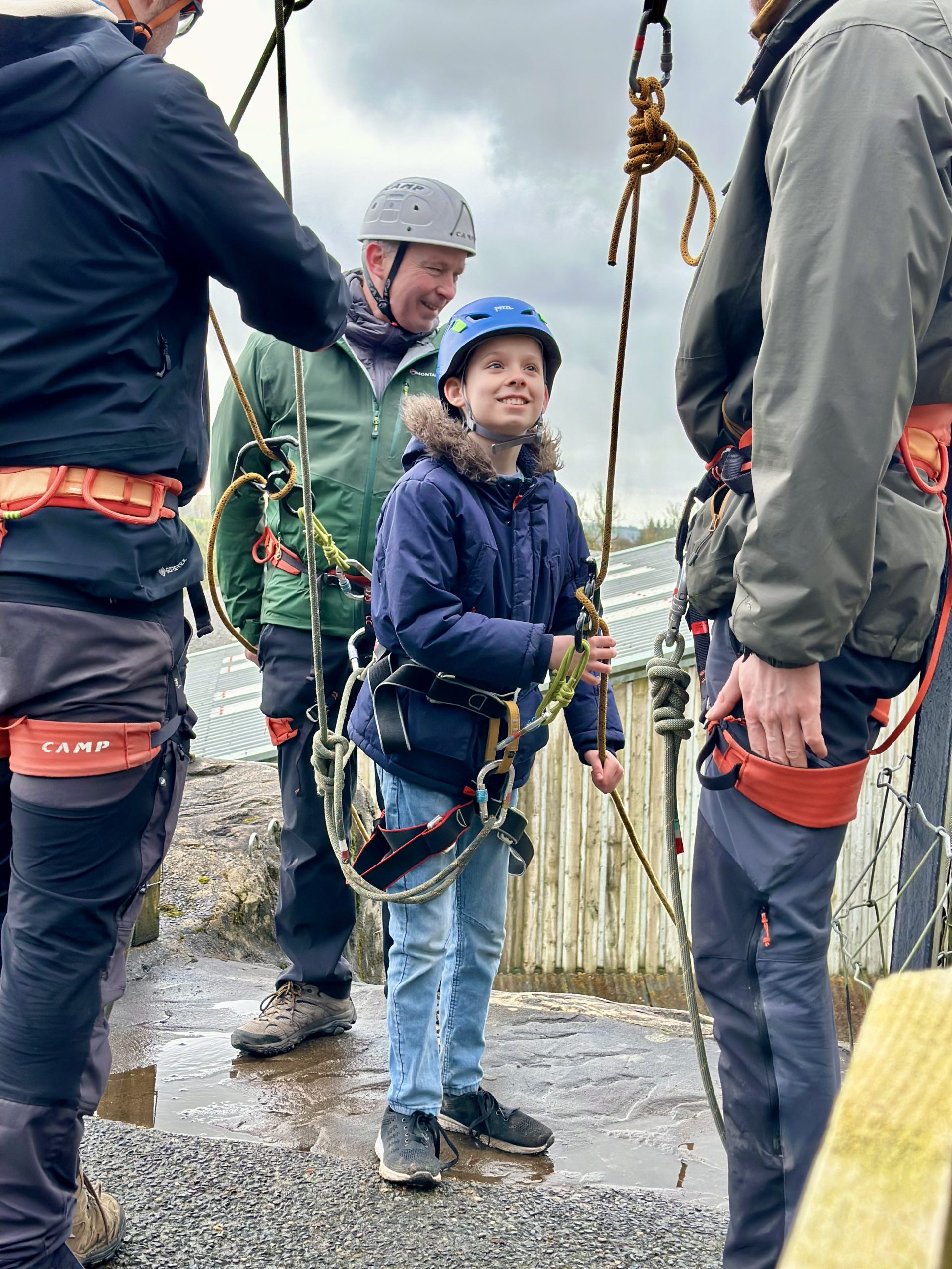 A young boy in a blue coat and a blue helmet is looking upwards and smiling at an adult standing in front of him. He is wearing a harness and has ropes attached to him as well as the person behind him, who is smiling.