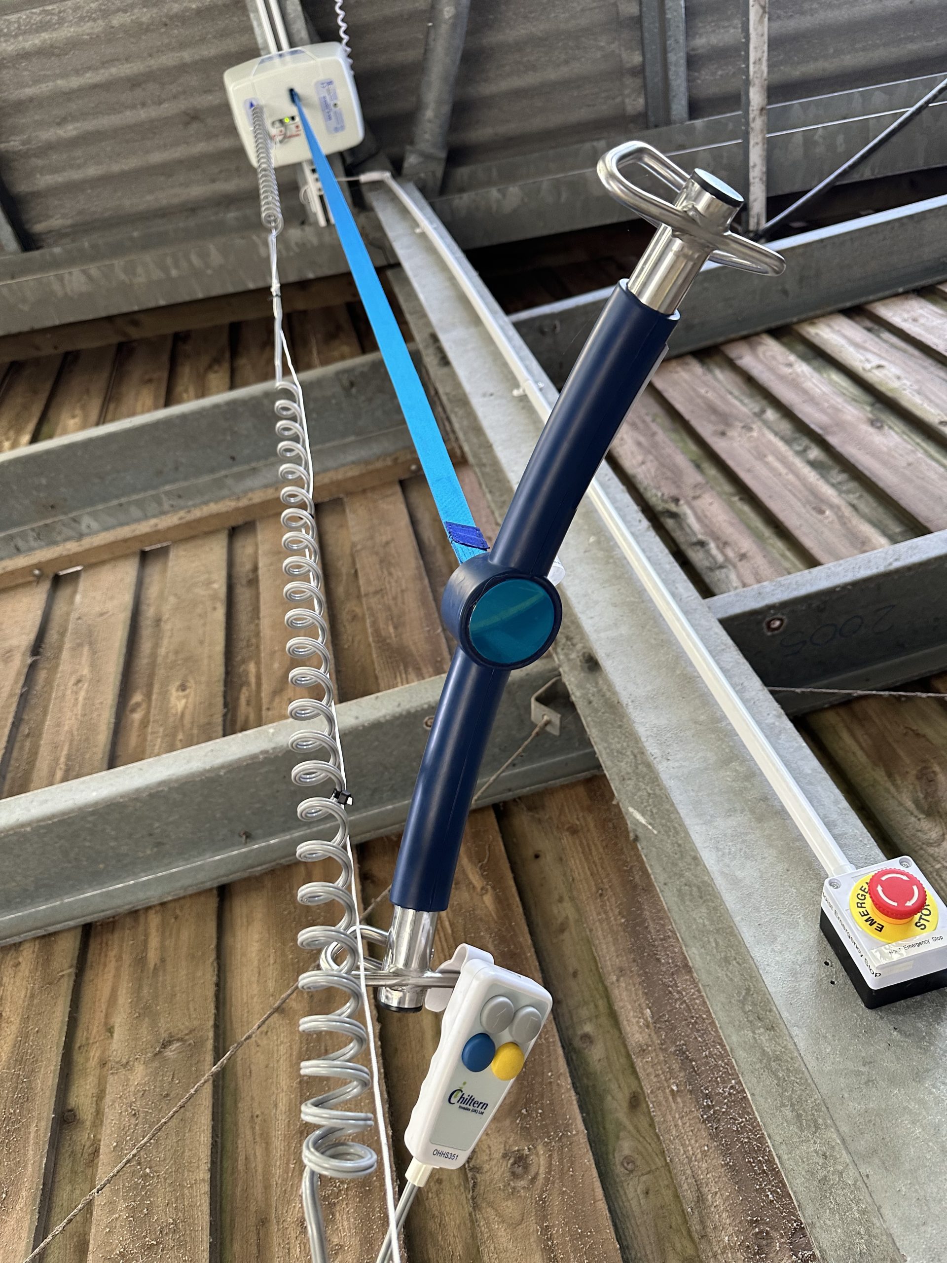 A view from below of a ceiling track hoist attached to a barn ceiling. It is a T-shaped blue bar with metal hooks at either end. There is a coiled wire hanging down alongside a blue belt attached to the bar. Attached to both is a remote control with coloured buttons.