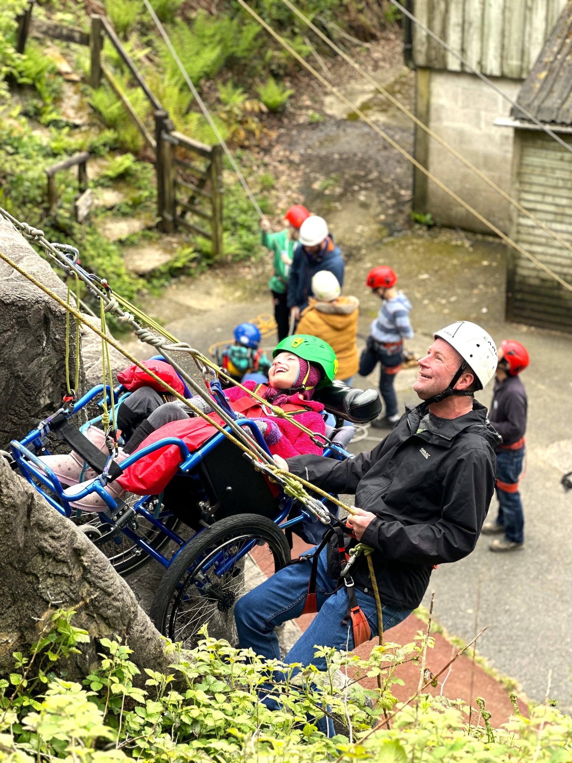 Two guests side by side on the abseil wall, one is in a wheelchair and the other is standing by her side and they are both smiling and looking up to the top of the wall. They are attached into a rope system and there are people blurred out in the background at the bottom of the climbing wall.