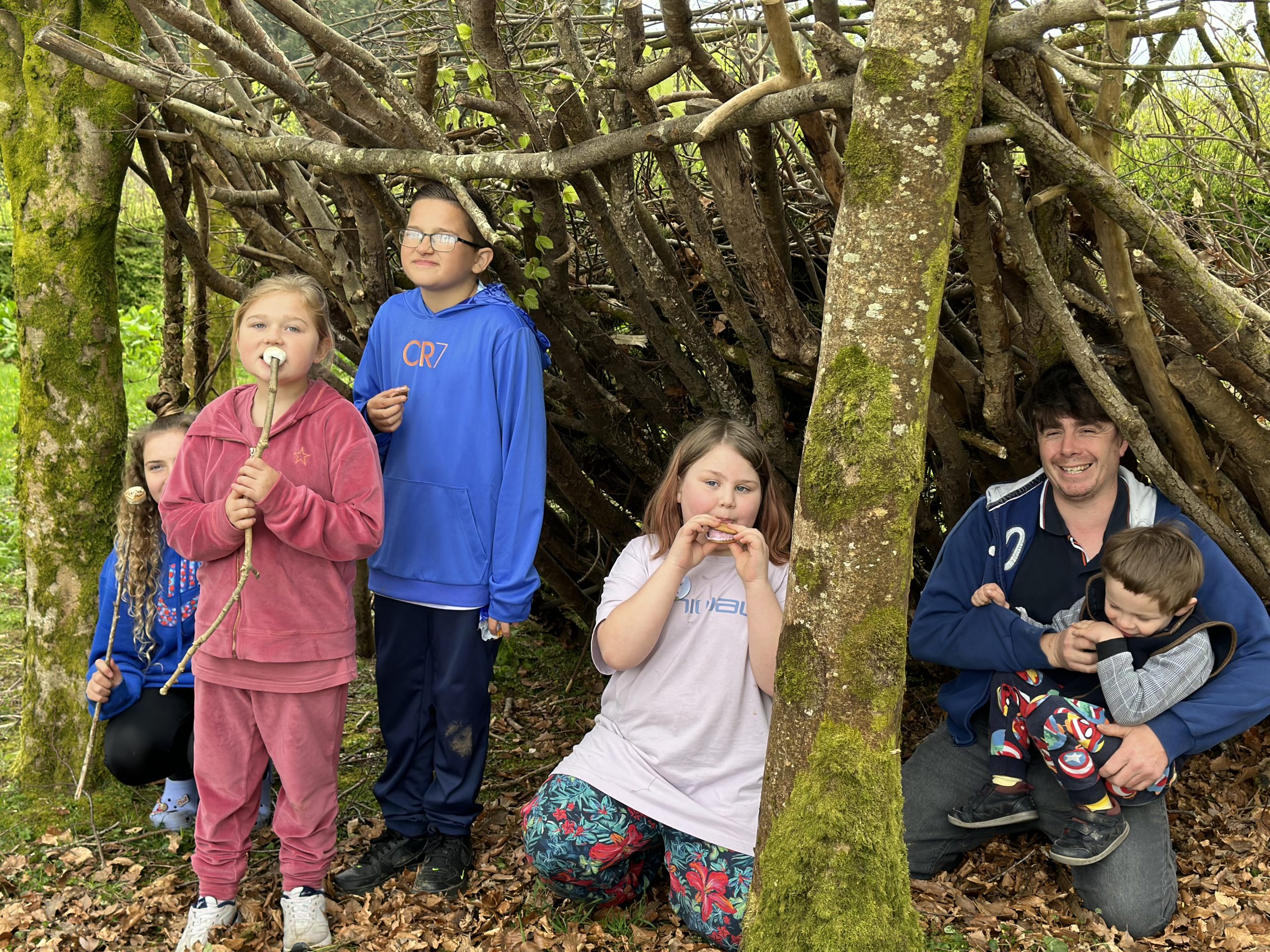 4 children and an adult holding a toddler are under a wooden-built shelter between two trees, smiling at the camera and eating marshmallows.