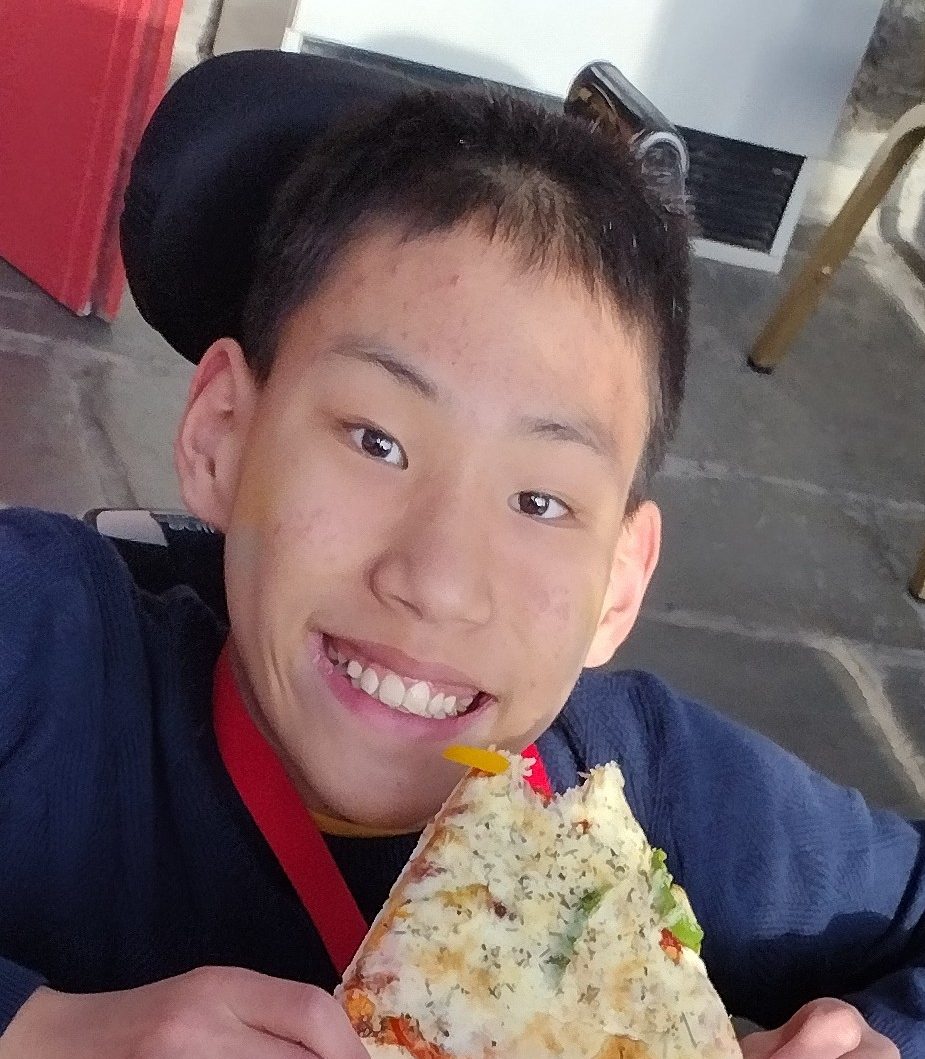 An image of a boy smiling at the camera, holding a slice of pizza in front of his face.